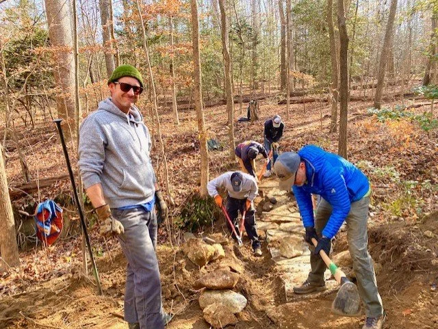 Group of people, including one man in a gray hoodie and green beanie, working on building a stone pathway in a wooded area during fall. They are using shovels and wearing gloves.