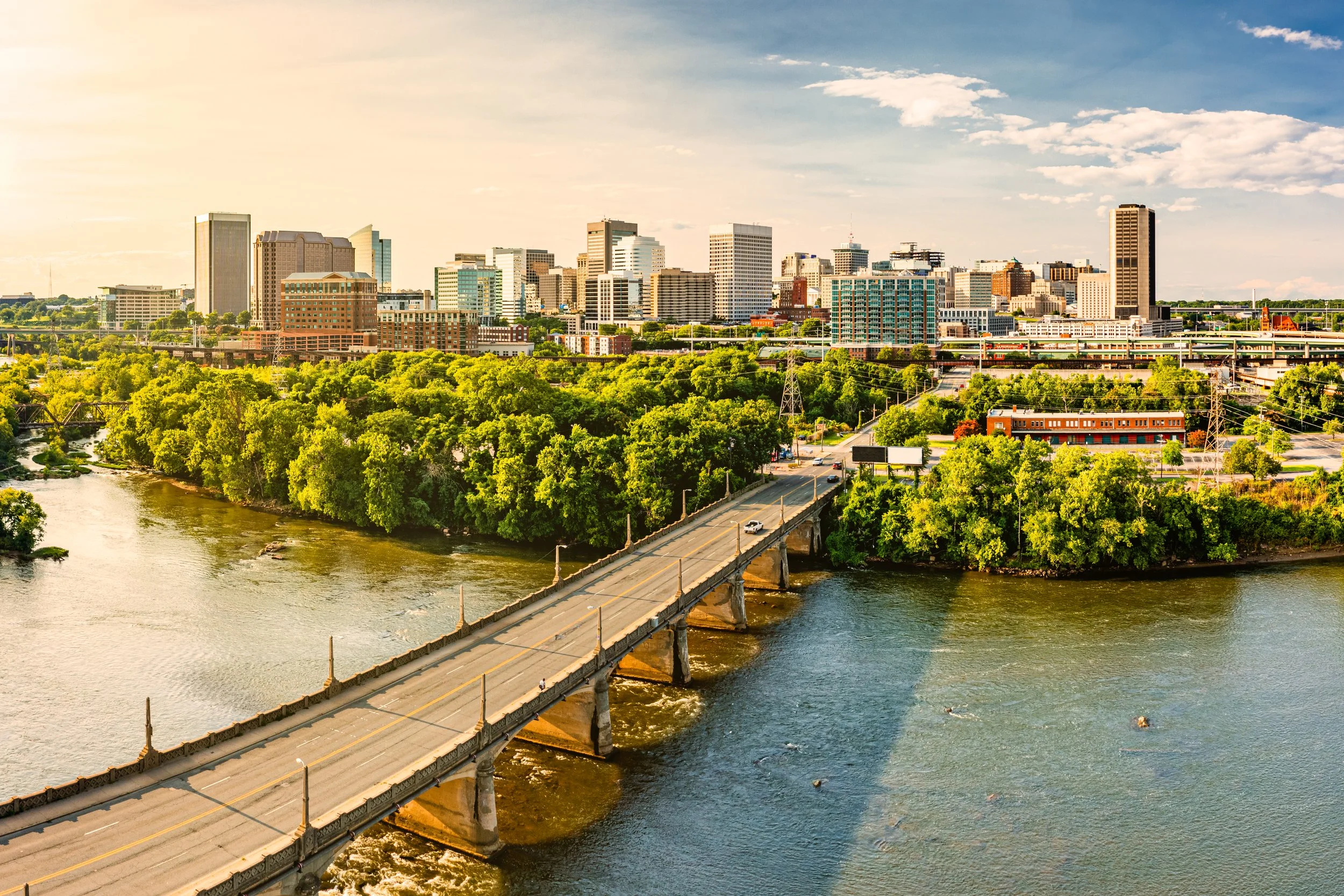 Aerial view of a city skyline with numerous buildings, a bridge crossing over a river, and green trees along the riverbank on a sunny day with a partly cloudy sky.