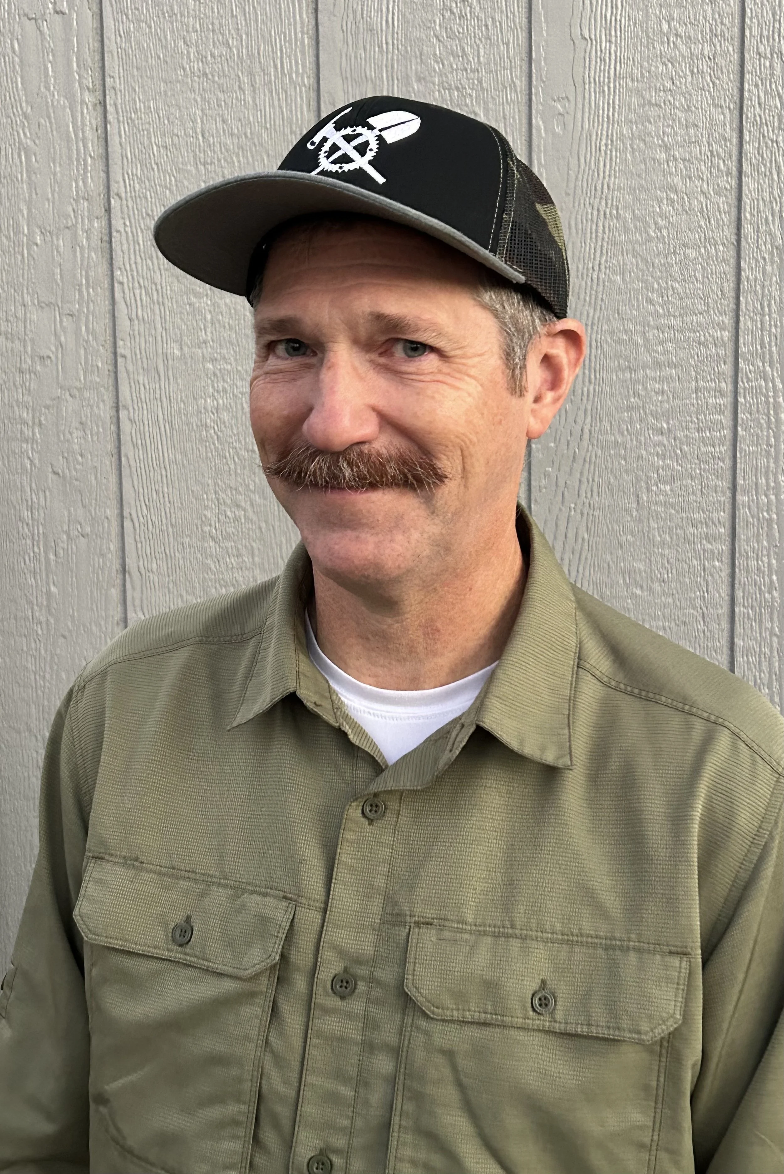 A man with a mustache and short hair wearing a black cap with a white design, a khaki button-up shirt, and a white undershirt stands in front of a gray wooden wall.