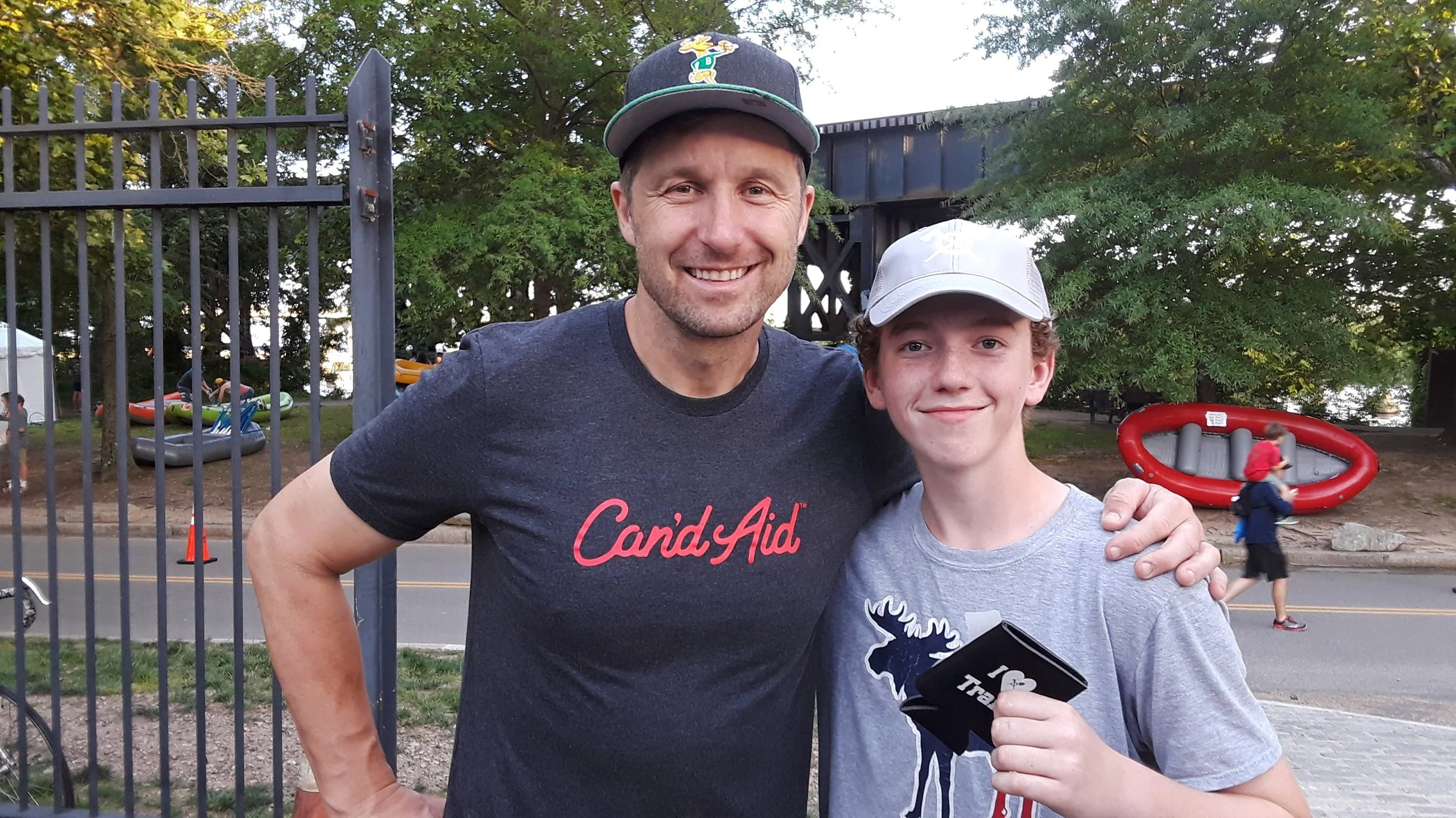 A man and a boy standing outdoors behind a metal fence, smiling at the camera. The man is wearing a dark gray T-shirt with 'Candy Aid' written on it and a dark baseball cap. The boy is wearing a light gray T-shirt and a white cap, holding a black item. In the background, there are trees, a road, and people playing or walking near an inflatable boat and other inflatable items.