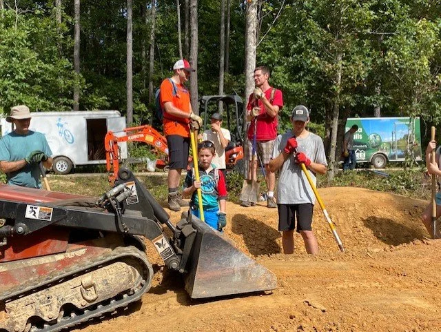 People working on a construction site in a forested area, with a small bulldozer and trucks in the background.
