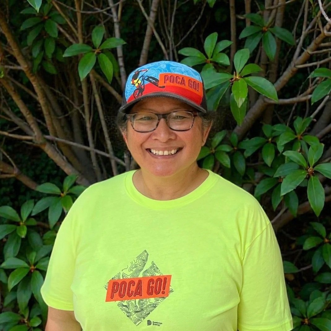A smiling woman with glasses, wearing a bright yellow T-shirt and a colorful cap, standing in front of green leafy bushes.