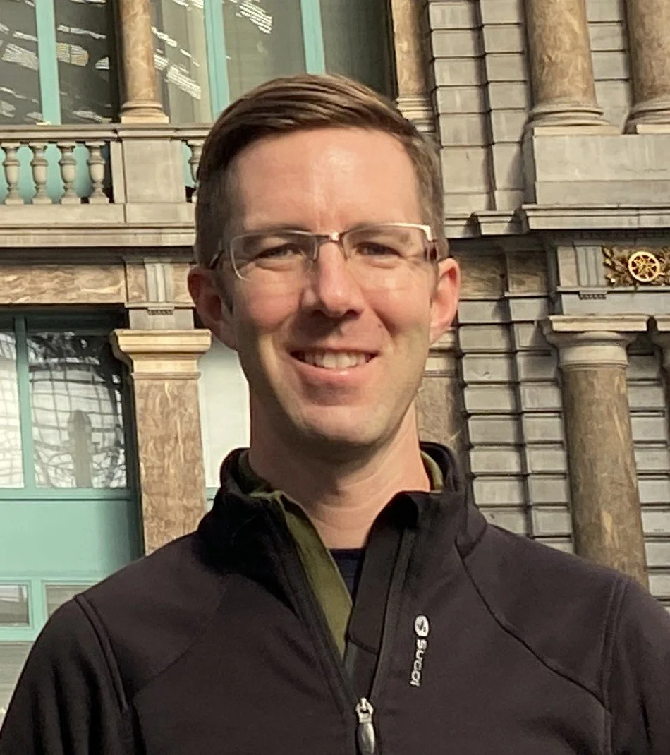 A man with glasses smiling outdoors in front of a historical stone building with large windows.