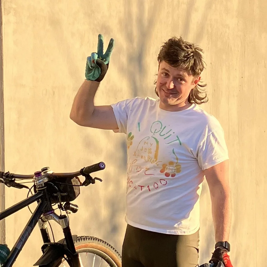 A man with short, dark, wavy hair smiling and making a peace sign with his left hand while standing next to a mountain bike, wearing a white t-shirt and gloves, against a plain, light-colored background.