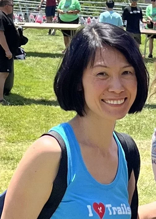 Smiling woman with shoulder-length black hair wearing a blue athletic shirt and carrying a backpack outdoors on a sunny day, with people in the background standing by tables.