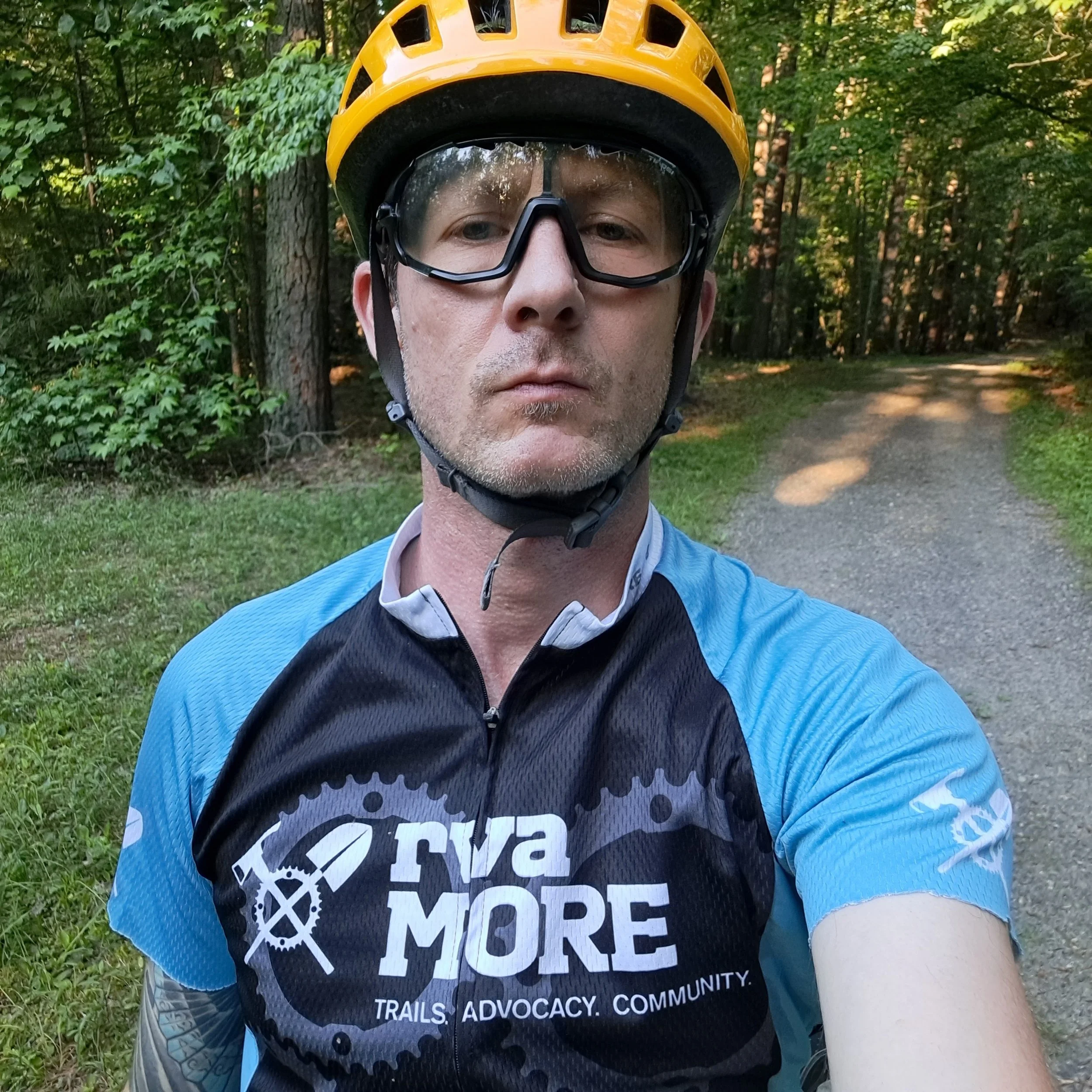 A man wearing a yellow bicycle helmet and black protective eyewear taking a selfie on a forest trail. He is dressed in a black and blue cycling jersey with the words 'SAVE MORE' and smaller text 'Trails. Advocacy. Community.' printed on it.