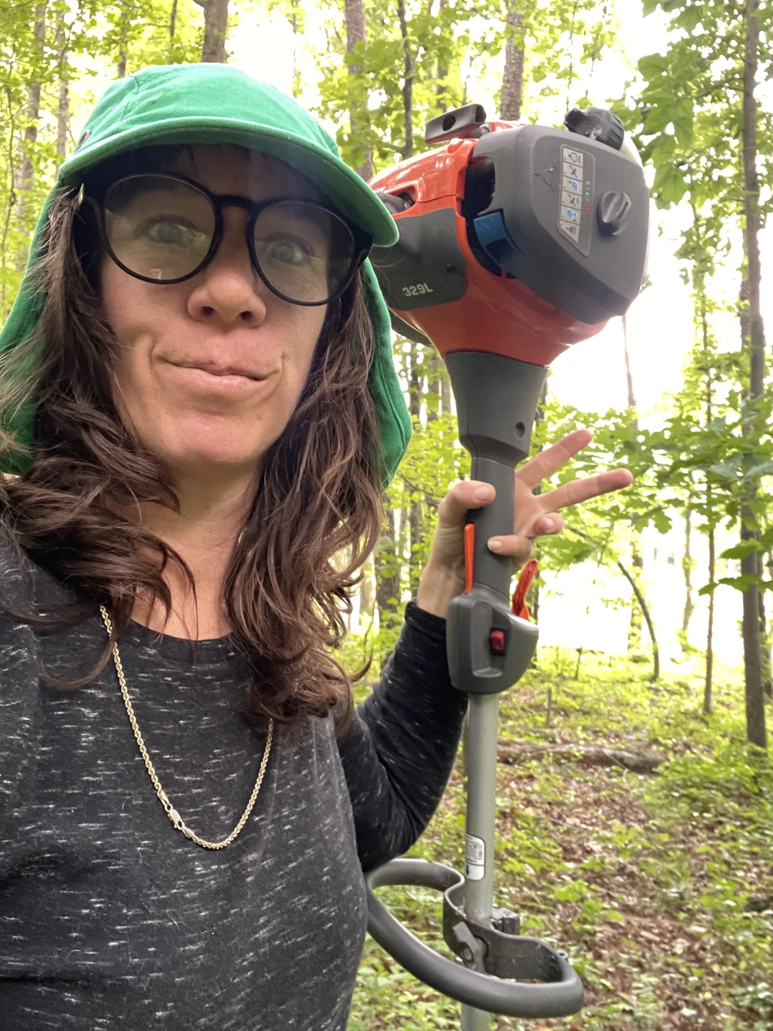 A woman wearing glasses, a green hat, and a black shirt standing in a forest, holding a leaf blower.