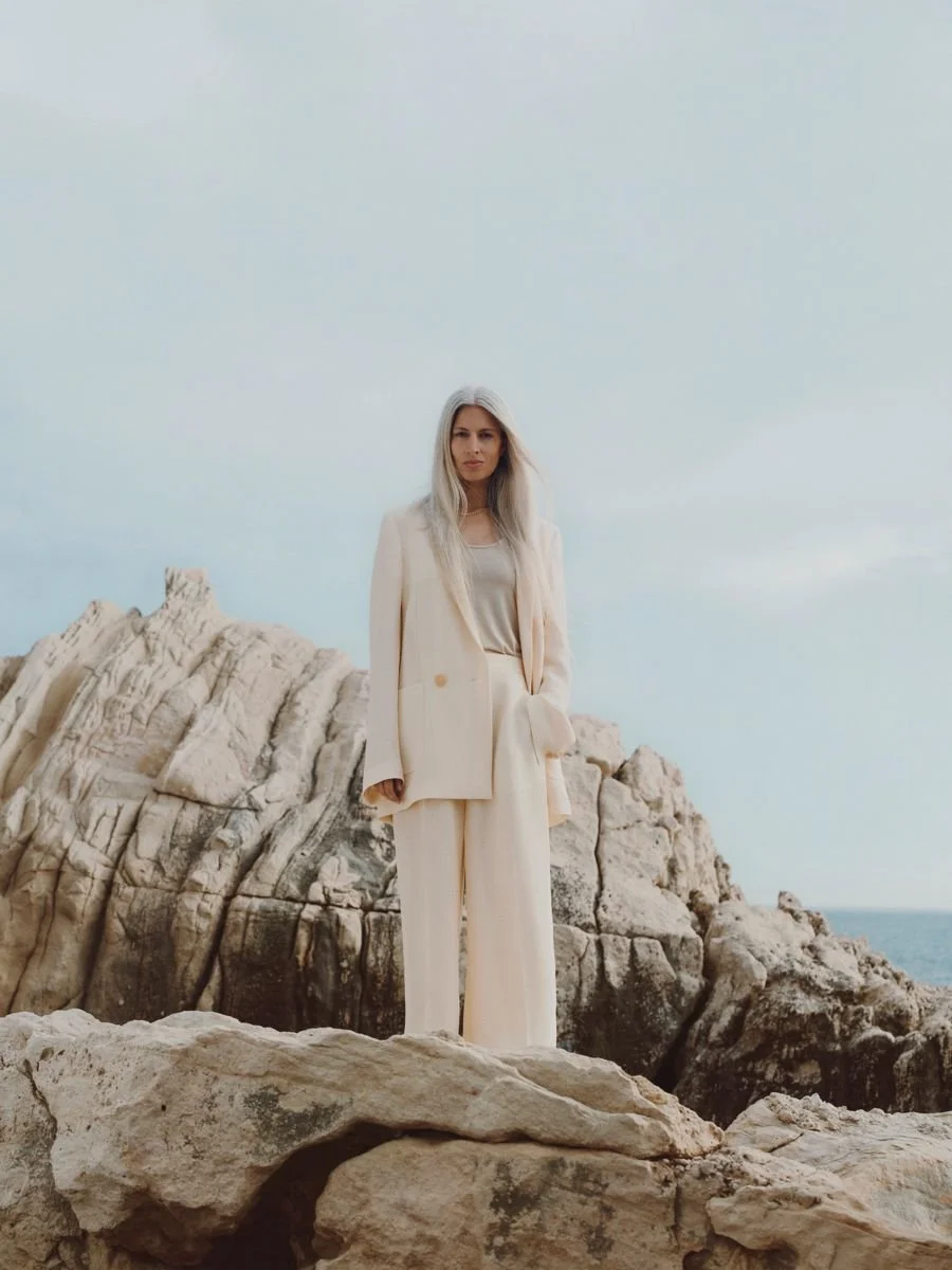 Femme portant un tailleur beige clair debout sur des rochers près de la mer, ciel nuageux en arrière-plan.