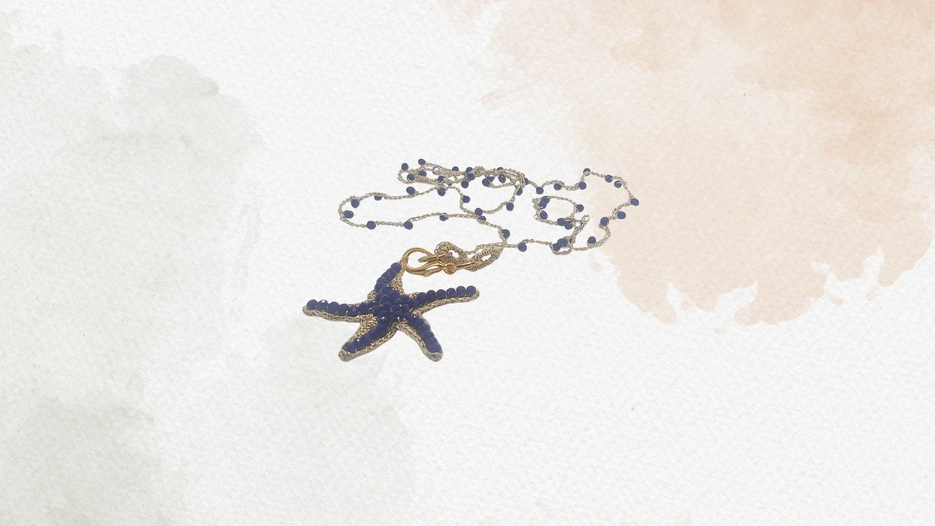 Una collana con pendente a forma di stelle marina blu con dettagli dorati, su uno sfondo bianco con un'ombra leggermente visibile.