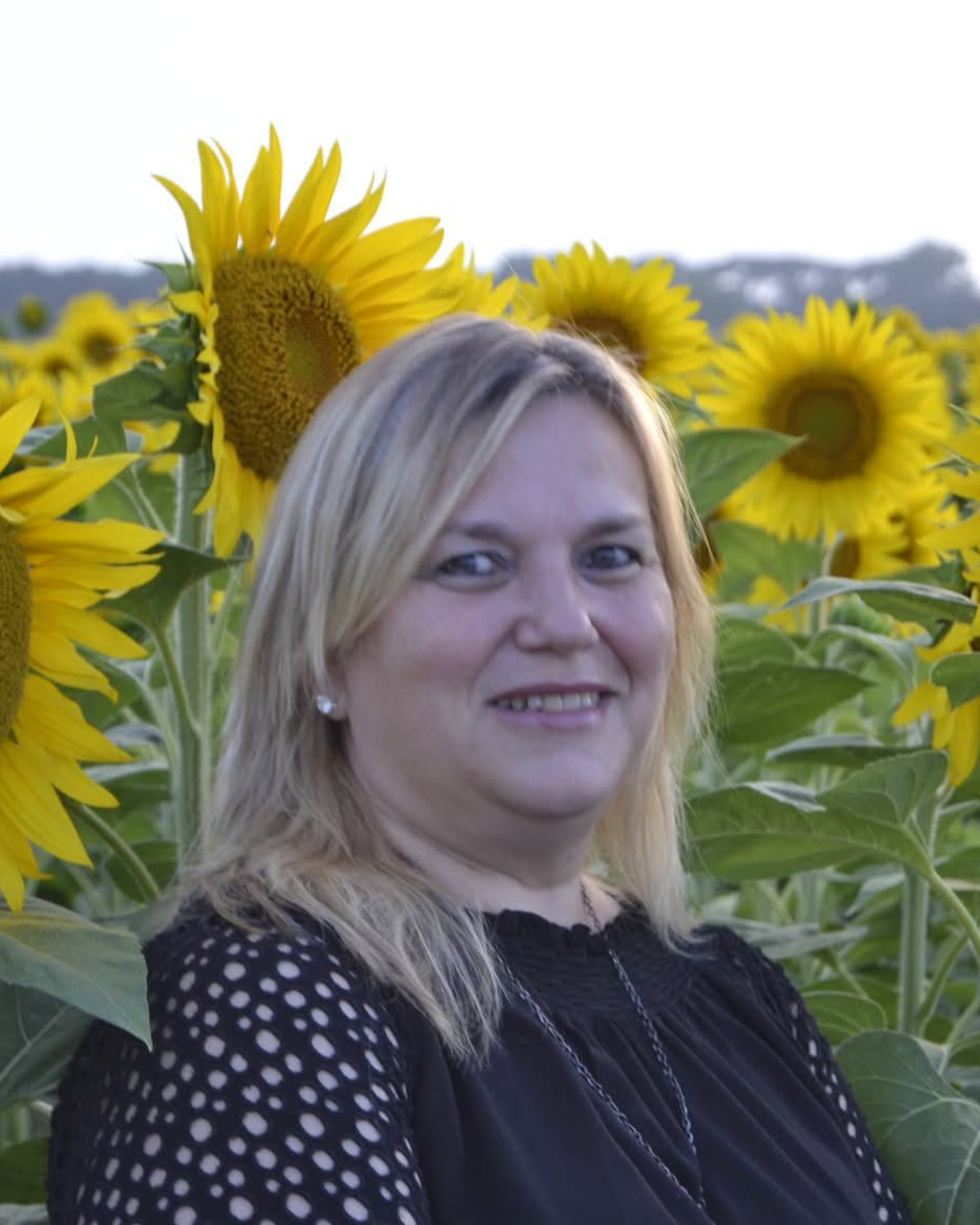 Una donna sorridente con capelli biondi in un campo di girasoli sotto un cielo chiaro.