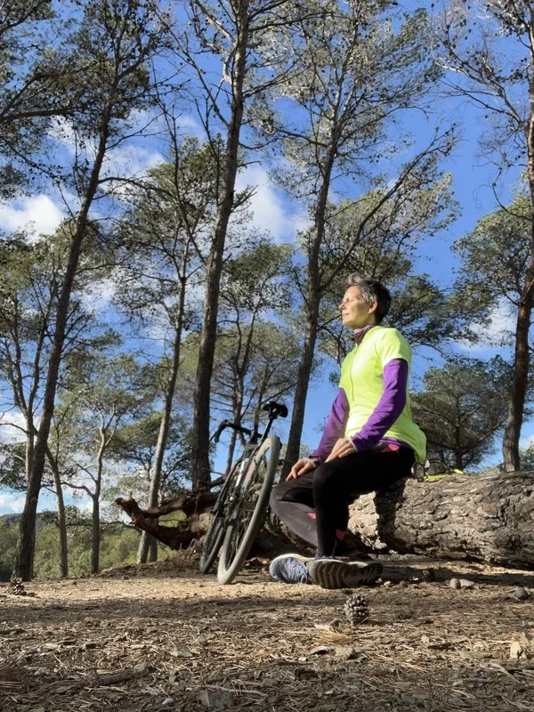 A woman sitting on a log in a forested area. She is wearing a bright yellow and purple jacket, black pants, and trail running shoes. A bicycle is leaning against the log next to her, and tall trees with sparse leaves are visible against a partly cloudy sky in the background.