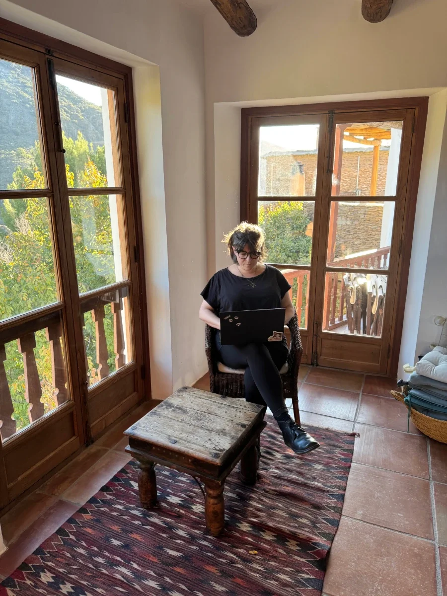 A woman with glasses sitting in a wicker chair, working on a laptop in a sunlit room with large wooden-framed windows and a view of trees and distant buildings outside.
