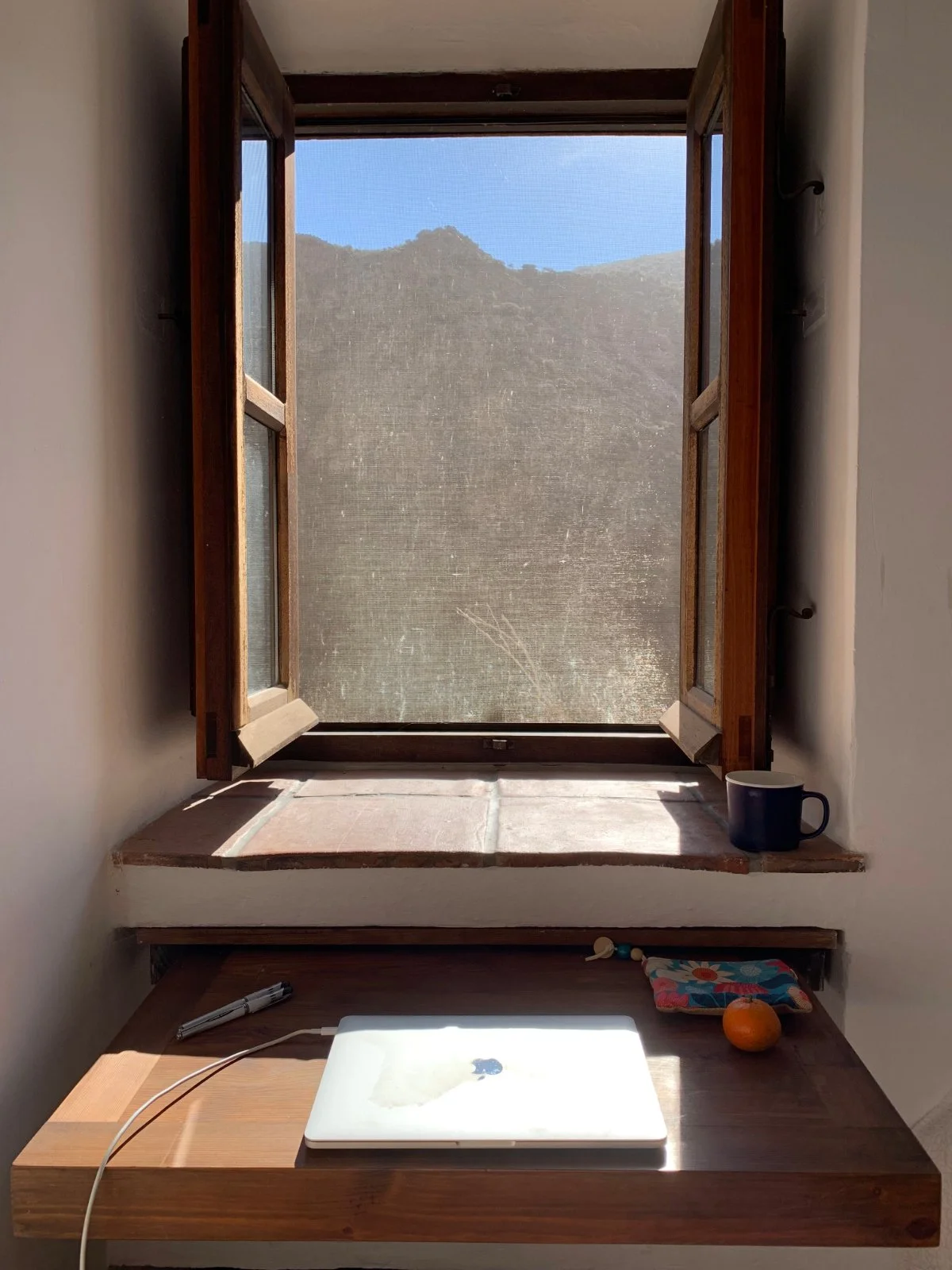 Open wooden window overlooking a mountainous landscape with blue sky, sunlight illuminating a small table with a closed Apple MacBook, a pen, a cup, a colorful cloth, an orange, and some small objects.