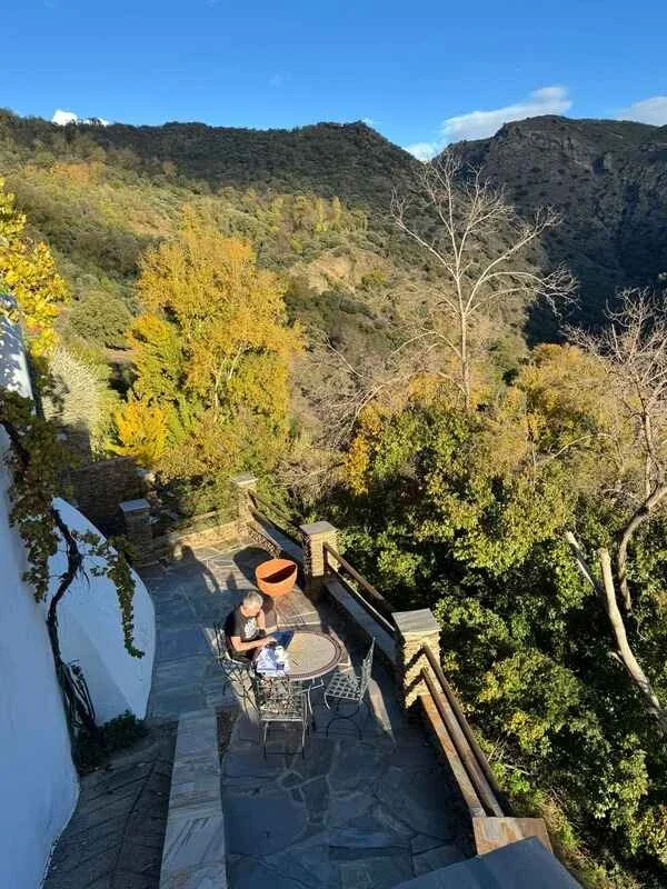 A person sitting at a round table on a balcony with a scenic view of mountains and trees, some leaves turning yellow and orange, with a clear blue sky in the background.