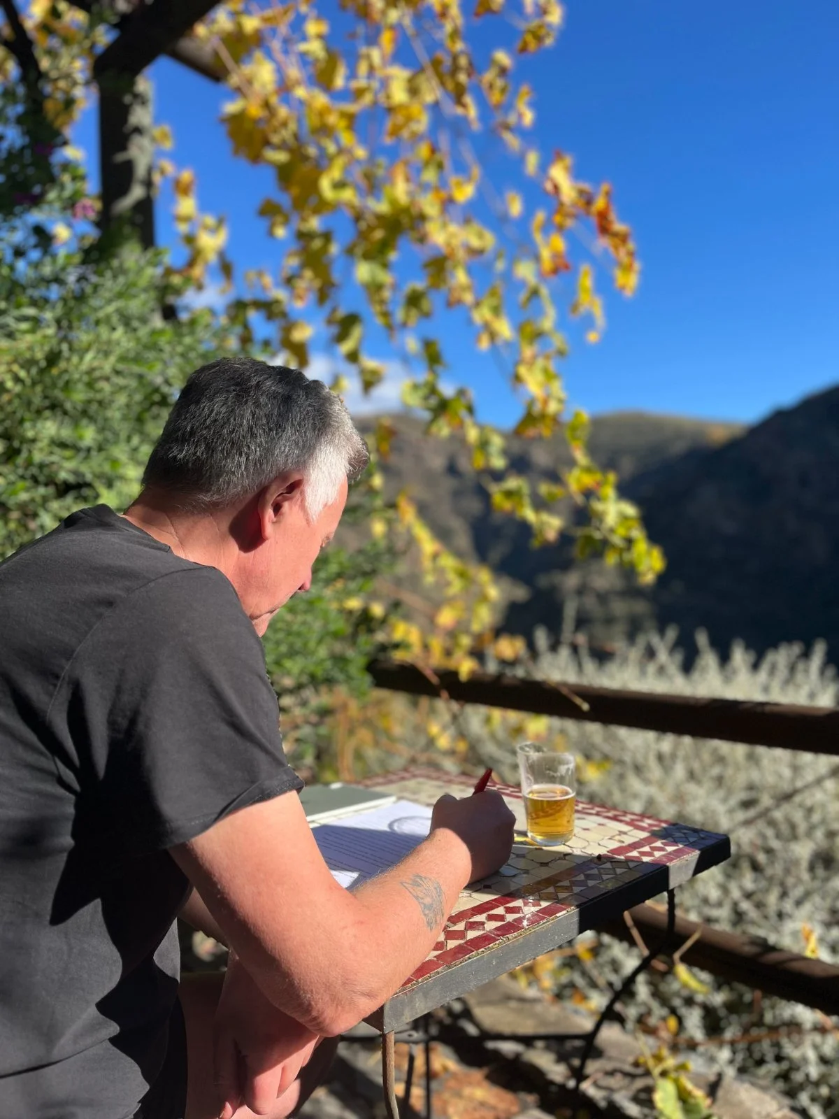 A man sitting at an outdoor table writing in a notebook with a glass of beer nearby, surrounded by trees with yellow leaves and mountains in the background.