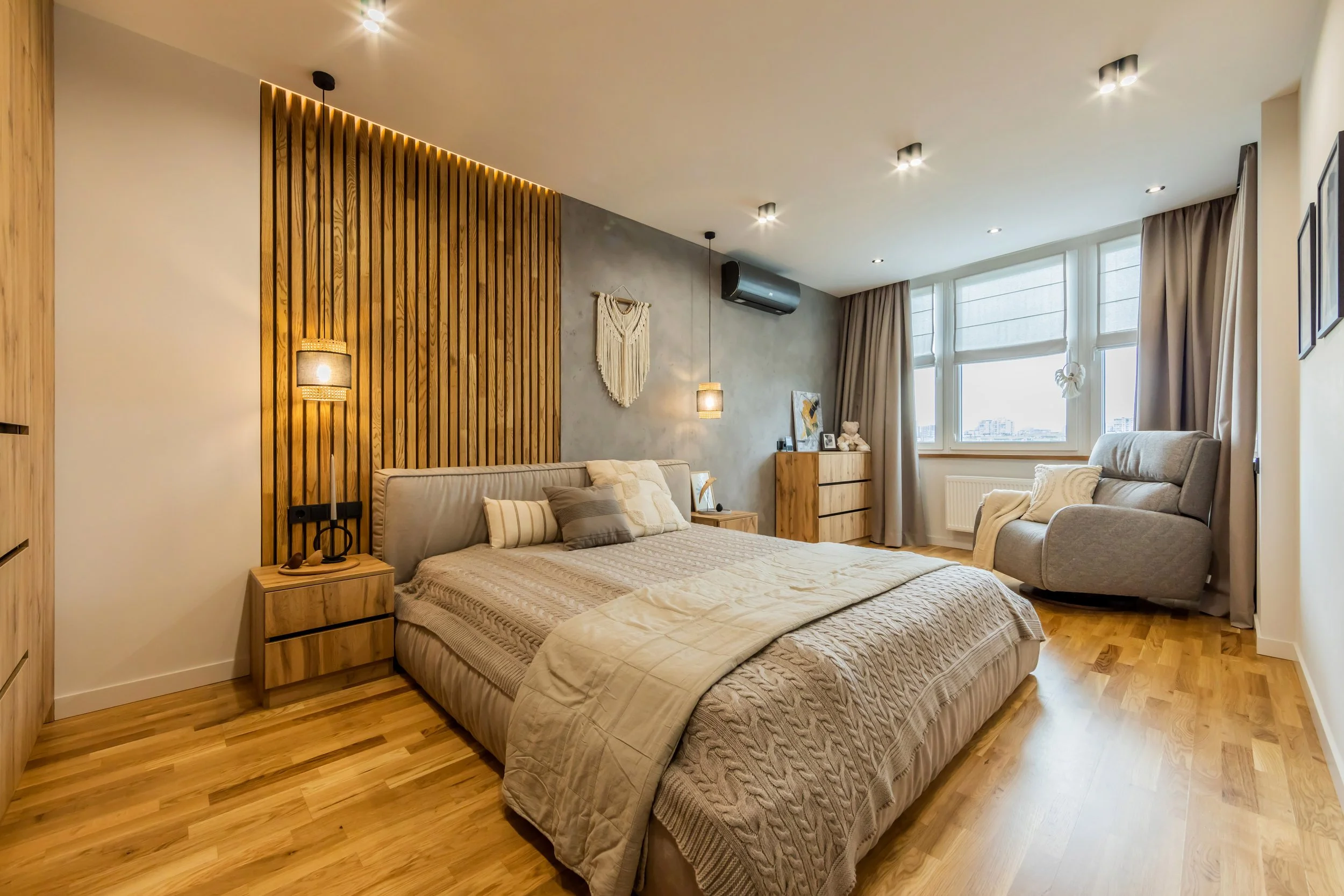 Modern bedroom with wood accents, cozy bed with pillows, bedside lamps, grey armchair near window, and wooden dresser.