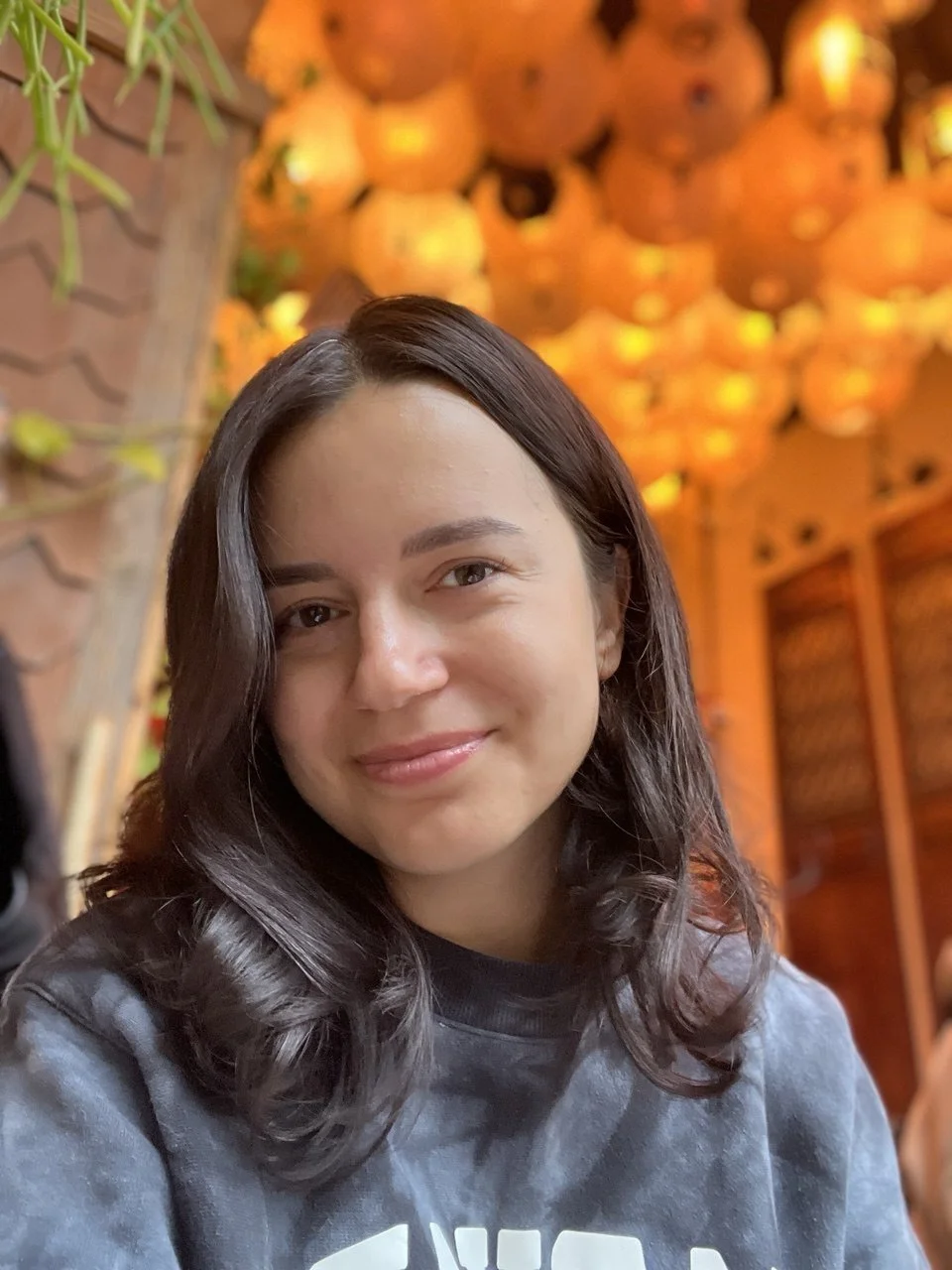 A young woman with dark hair smiling in a restaurant decorated with orange lanterns.