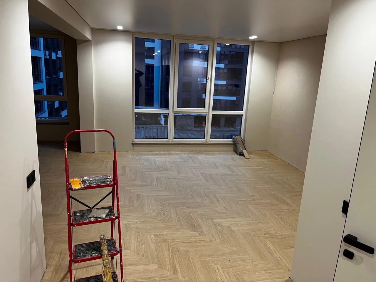 Empty room with light-colored wooden floor, large windows, and a red step ladder.