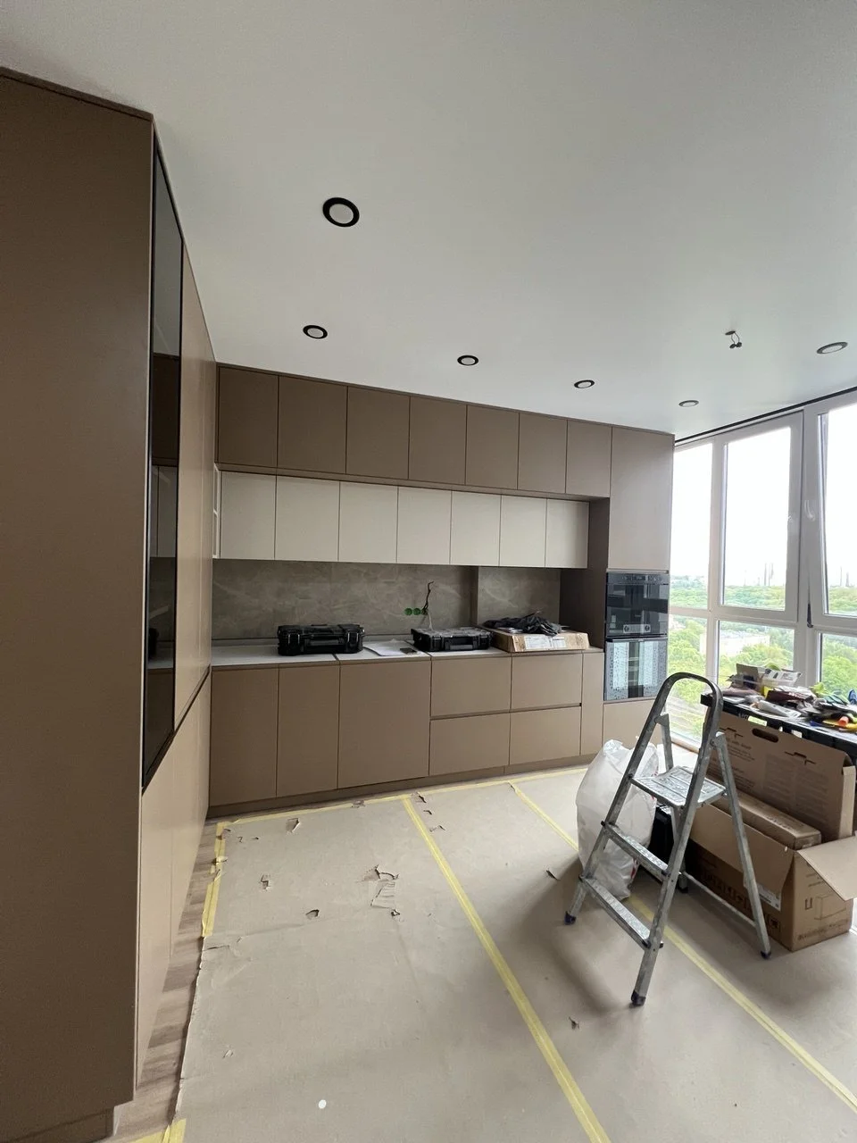 Kitchen under renovation with unfinished cabinets, tools, a ladder, and cardboard boxes near a large window.