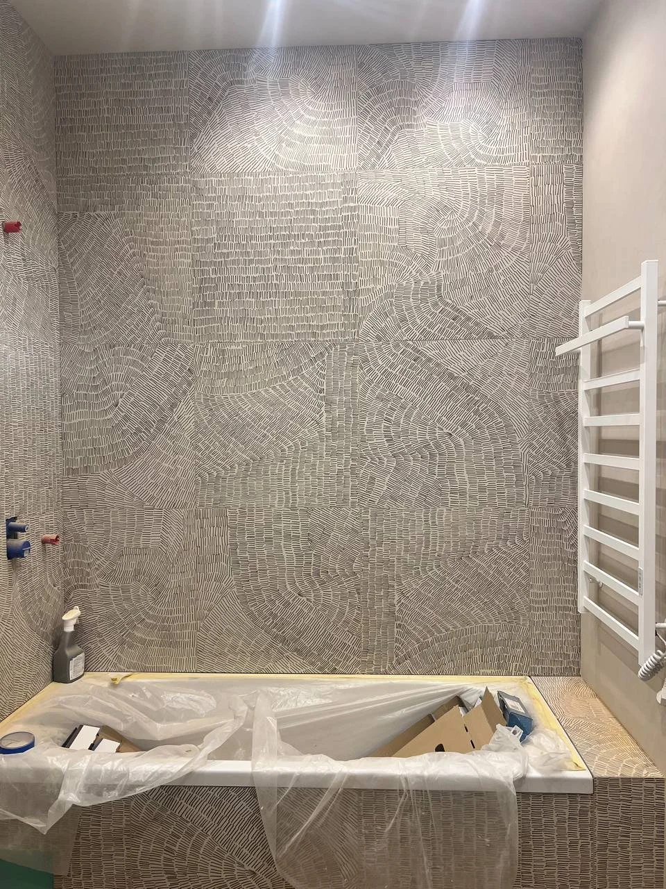 Bathroom under construction with a large, intricately patterned tile wall, a bathtub wrapped in plastic with construction materials inside, and a white towel rack on the right wall.