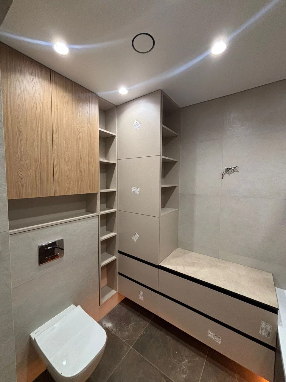 Bathroom with beige and wooden cabinets, a toilet, and a wall with a faucet and tiles.