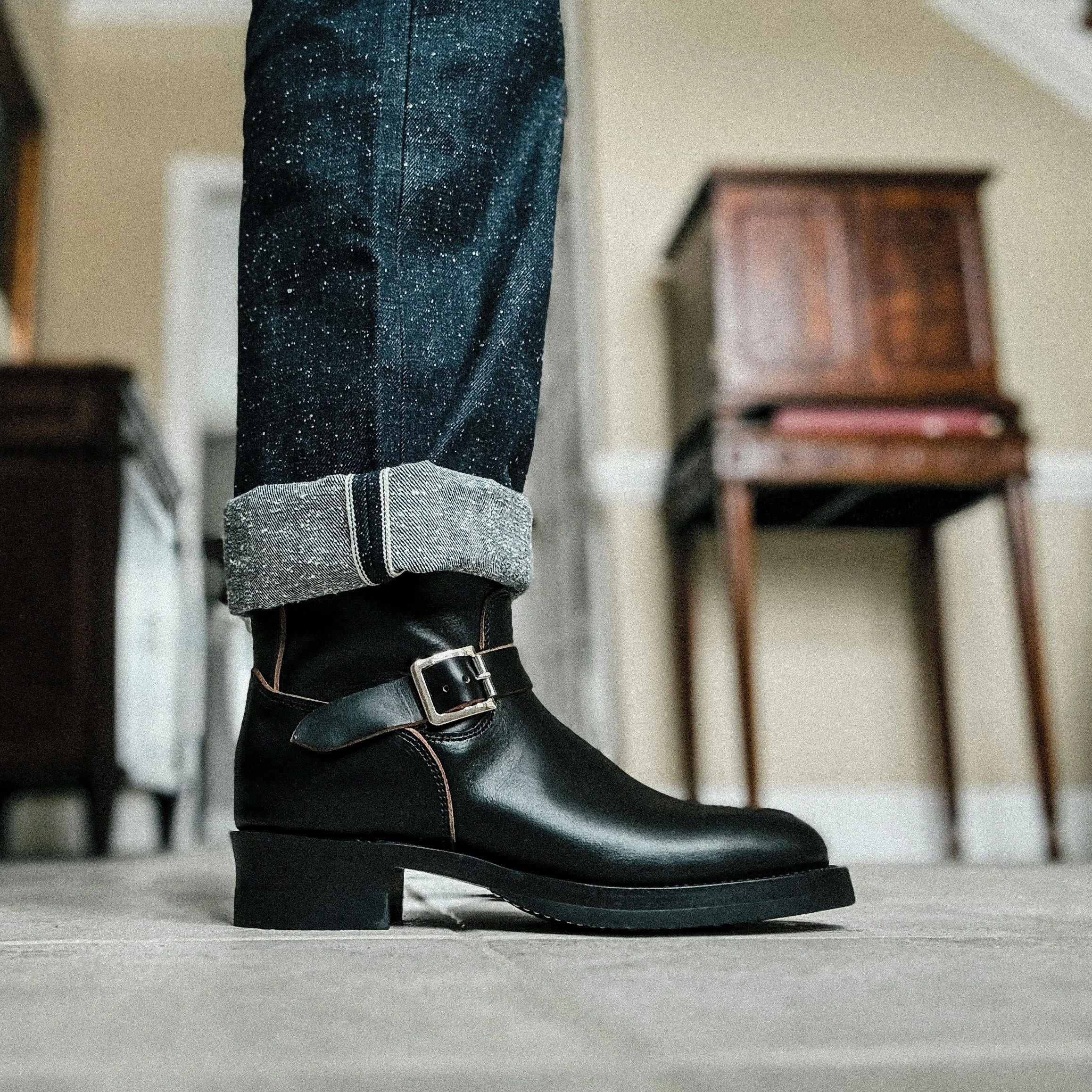 Close-up of a person wearing black leather boots with buckle detail, cuffed jeans with a speckled pattern, standing indoors near a wooden table.