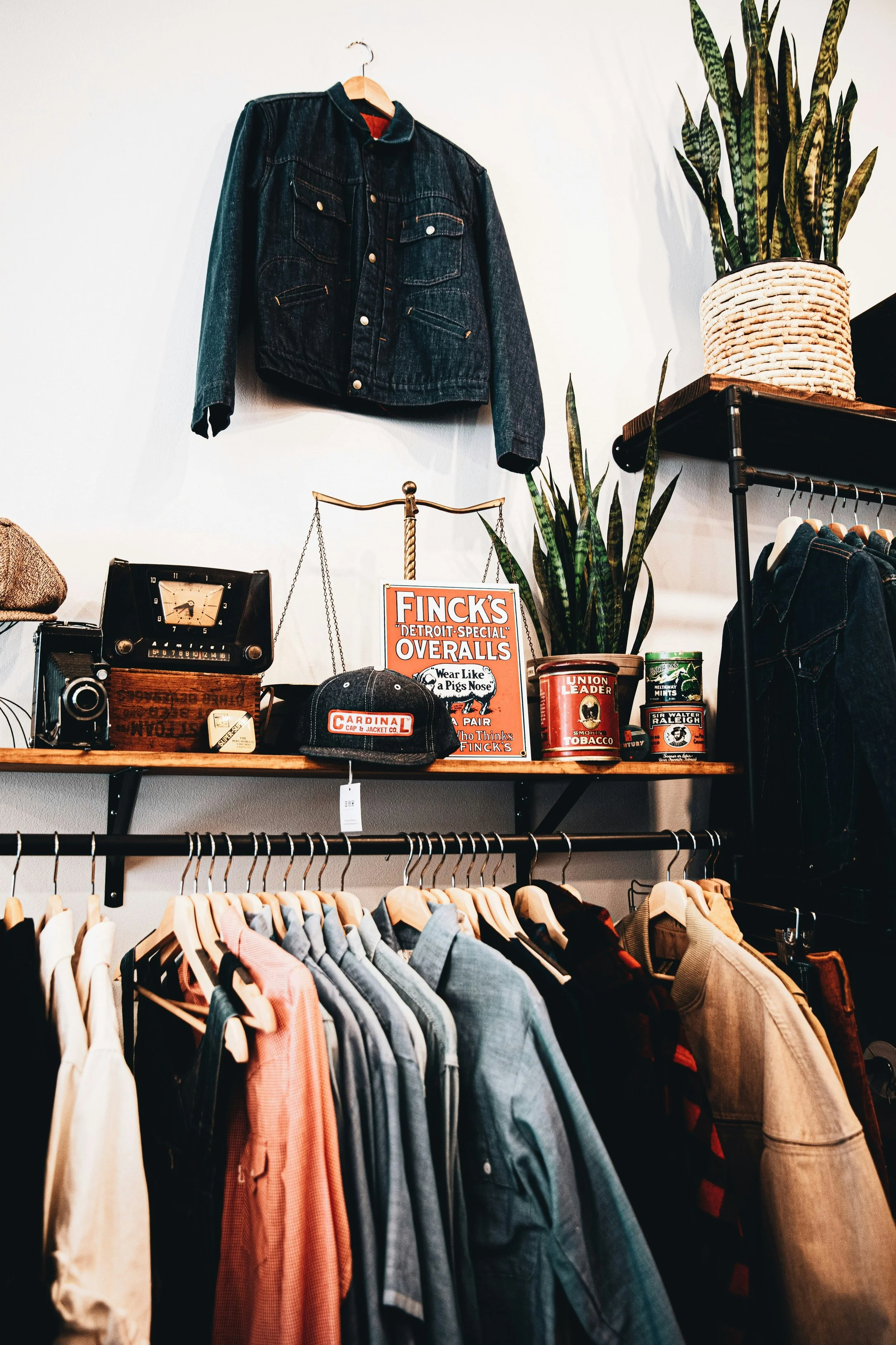 Display of vintage clothing and accessories in a boutique, including denim jackets, shirts, hats, signs, and plants.
