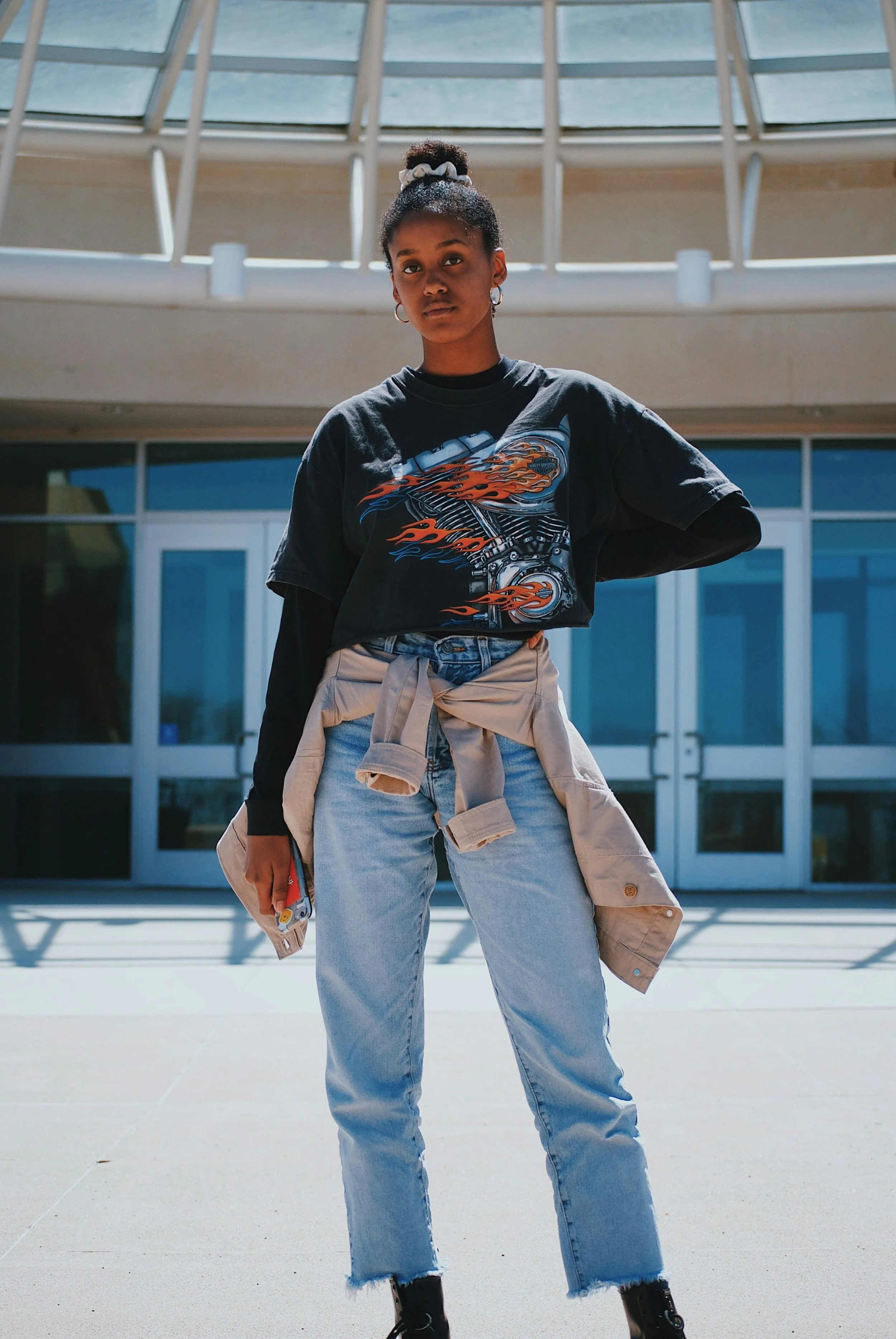 A young woman with a high ponytail, earrings, and medium skin, standing outdoors in front of a modern glass building, wearing a black graphic t-shirt, light-wash jeans with frayed hems, and a beige jacket tied around her waist.