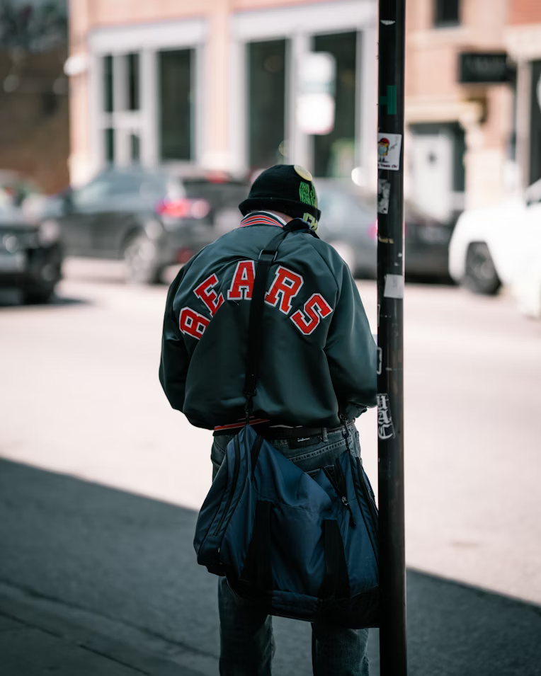 A person standing on a city sidewalk near a pole, wearing a black beanie, a dark jacket with 'BEARS' written on the back, and carrying a blue duffel bag.