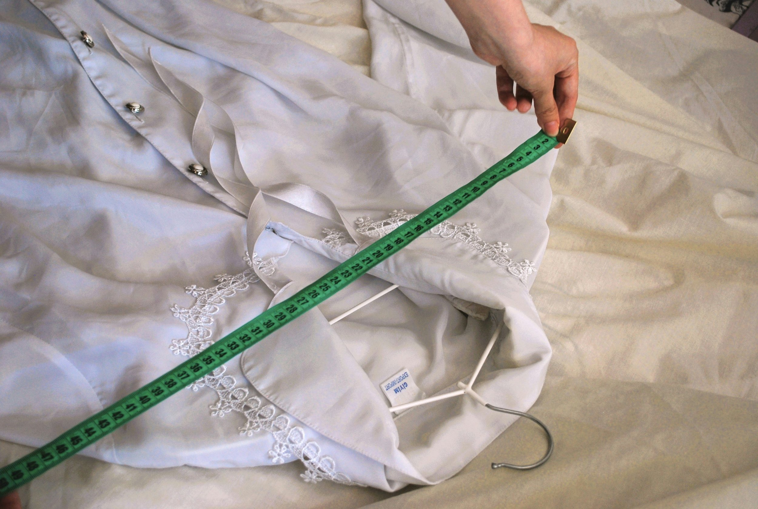 A person using a measuring tape to measure a white satin and lace bedspread or garment, with a white hanger beneath.