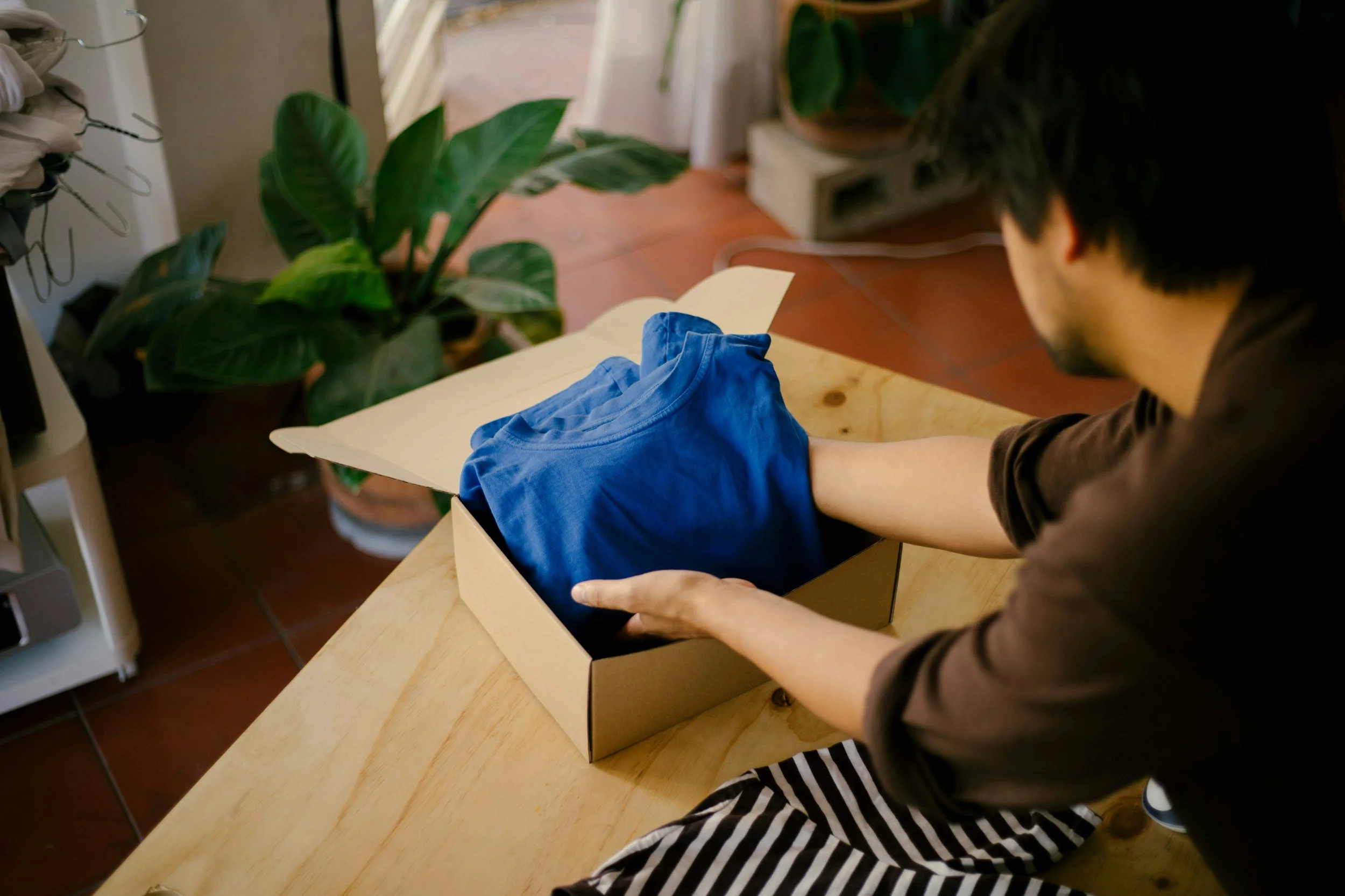 A person packing a blue t-shirt into a cardboard box, sitting at a wooden table in a room with potted plants and tiled flooring.