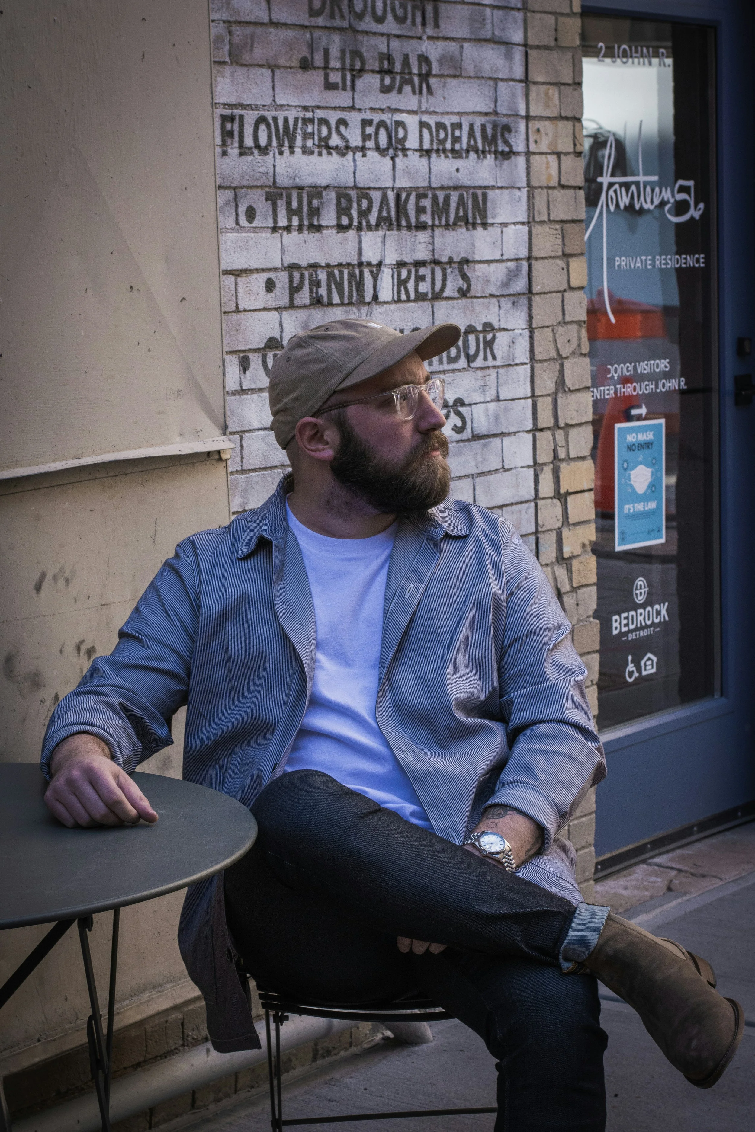 A bearded man wearing glasses, a beige cap, a light jacket over a white shirt, and dark jeans, is sitting outside on a chair beside a small round table near a brick building with signs and a window.