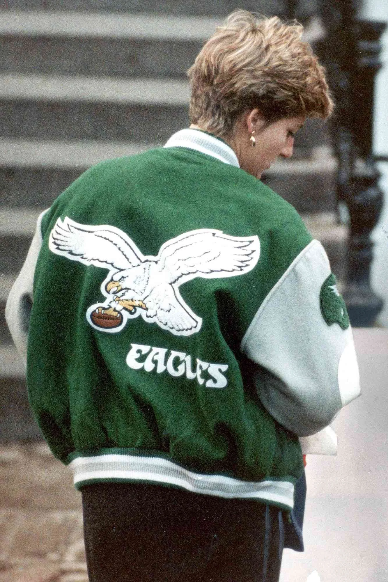 A person with short, wavy hair wearing a green and gray varsity jacket with a large eagle logo on the back and the word 'EAGLES' underneath, standing outdoors.