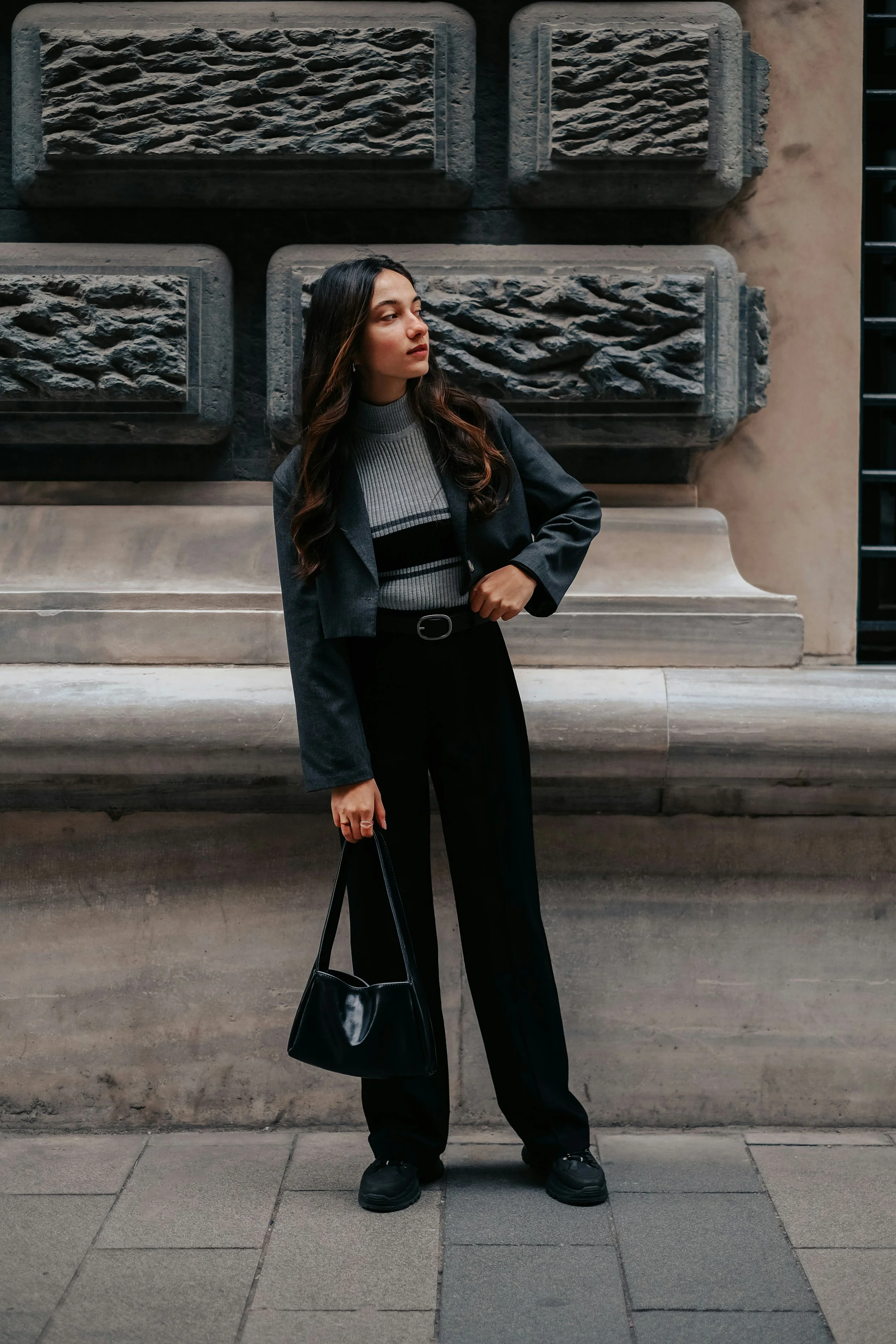 A young woman with long dark hair wearing a gray and black turtleneck sweater, black blazer, and black pants, standing on a city sidewalk in front of a textured stone wall.