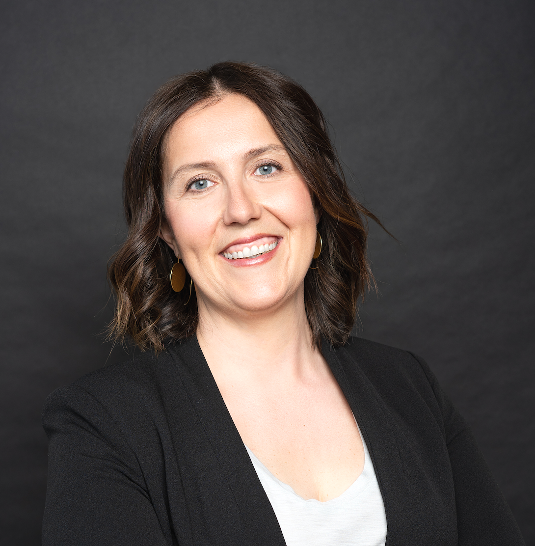 Professional woman with shoulder-length brown hair, wearing a black blazer and a white top, smiling against a dark gray background.