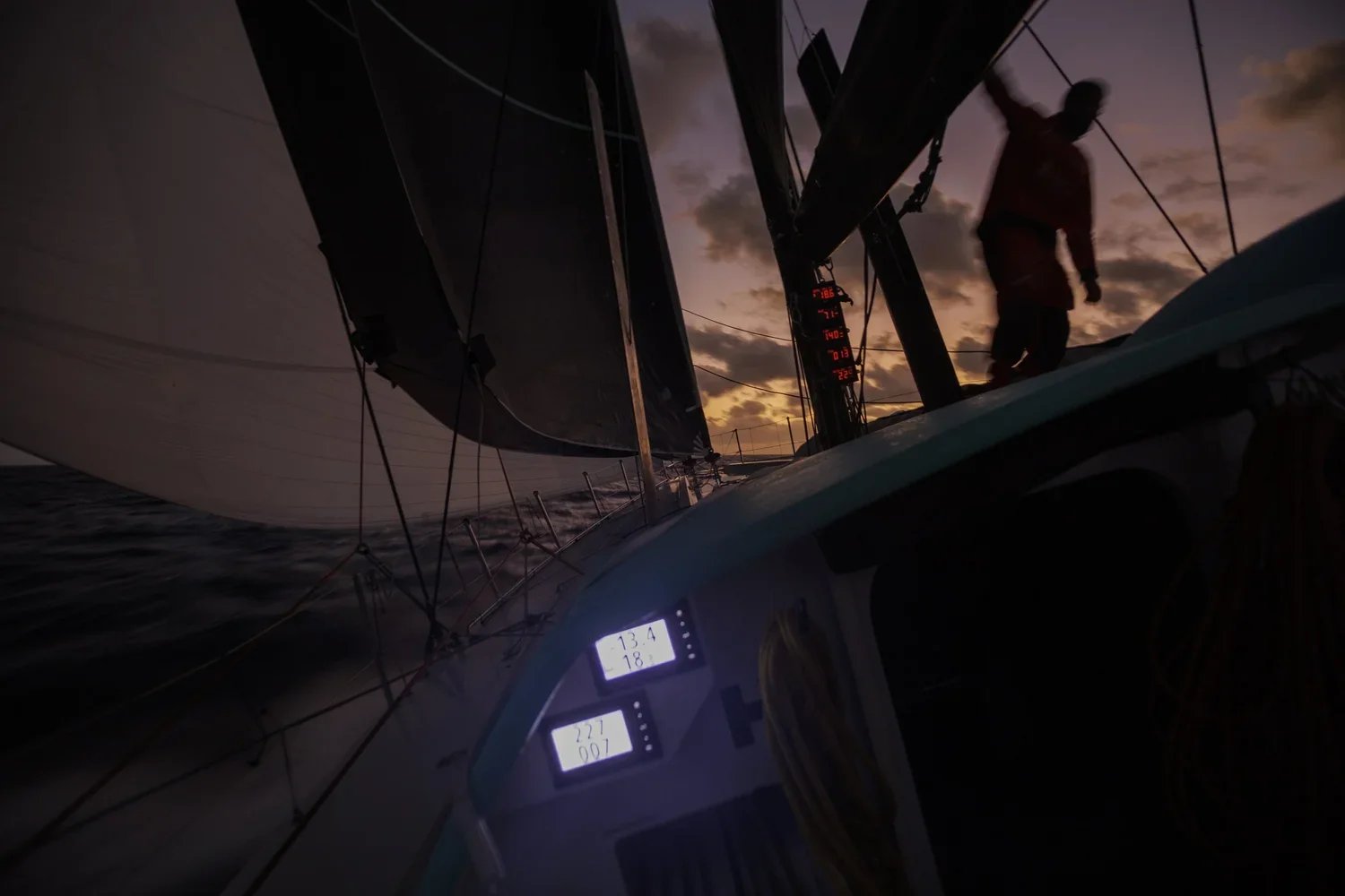 Une personne en navigation nocturne sur un voilier, avec un ciel au crépuscule, affichage électronique montrant des données de navigation, et les voiles déployés.