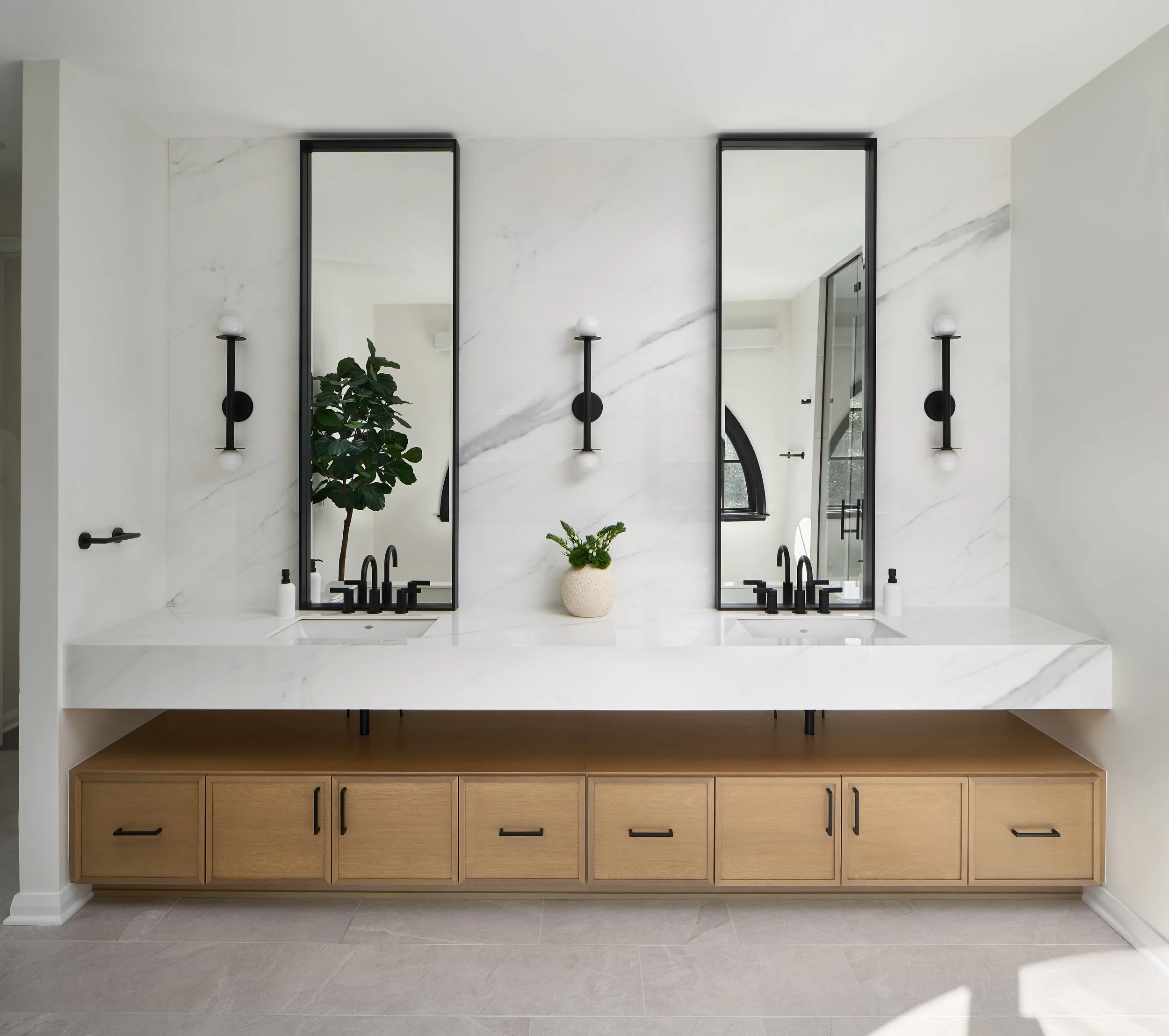A Luxury Modern bathroom renovation by Benham & Co Interior Design based in Columbus Ohio.  With white marble walls, black fixtures, a wooden cabinet, and decorative black wall sconces with white globes.