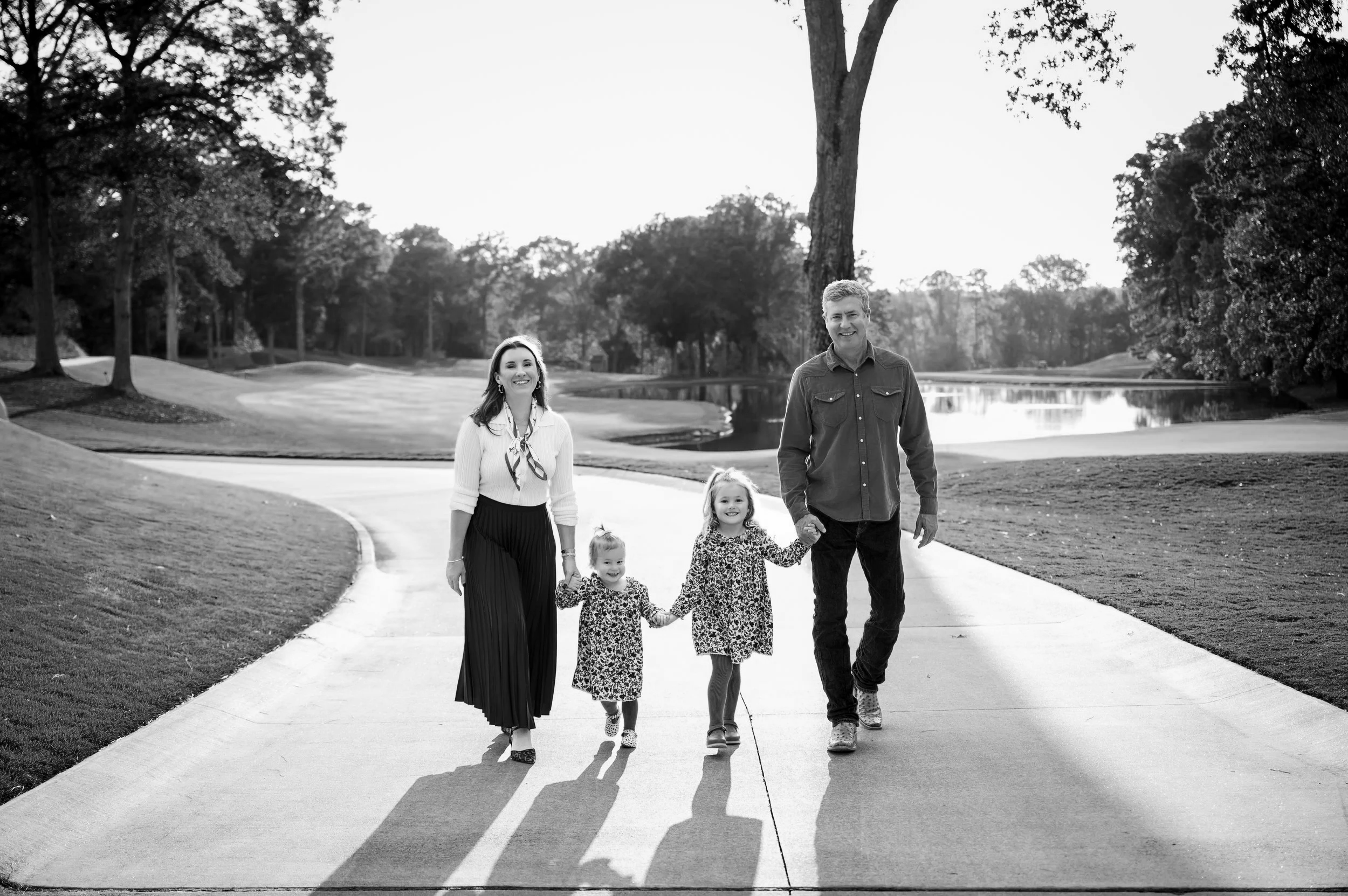 A black and white photo of a family of four walking on a park sidewalk, holding hands, with a lake and trees in the background.