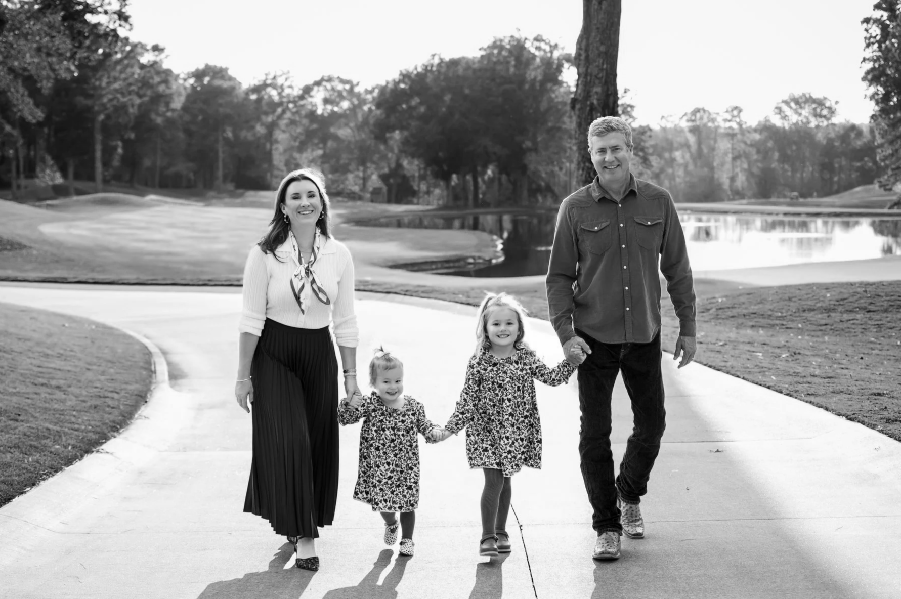 A family of four, including a woman, a man, and two young girls, walking hand-in-hand on a paved path near a pond in a park. They are smiling and enjoying a sunny day.