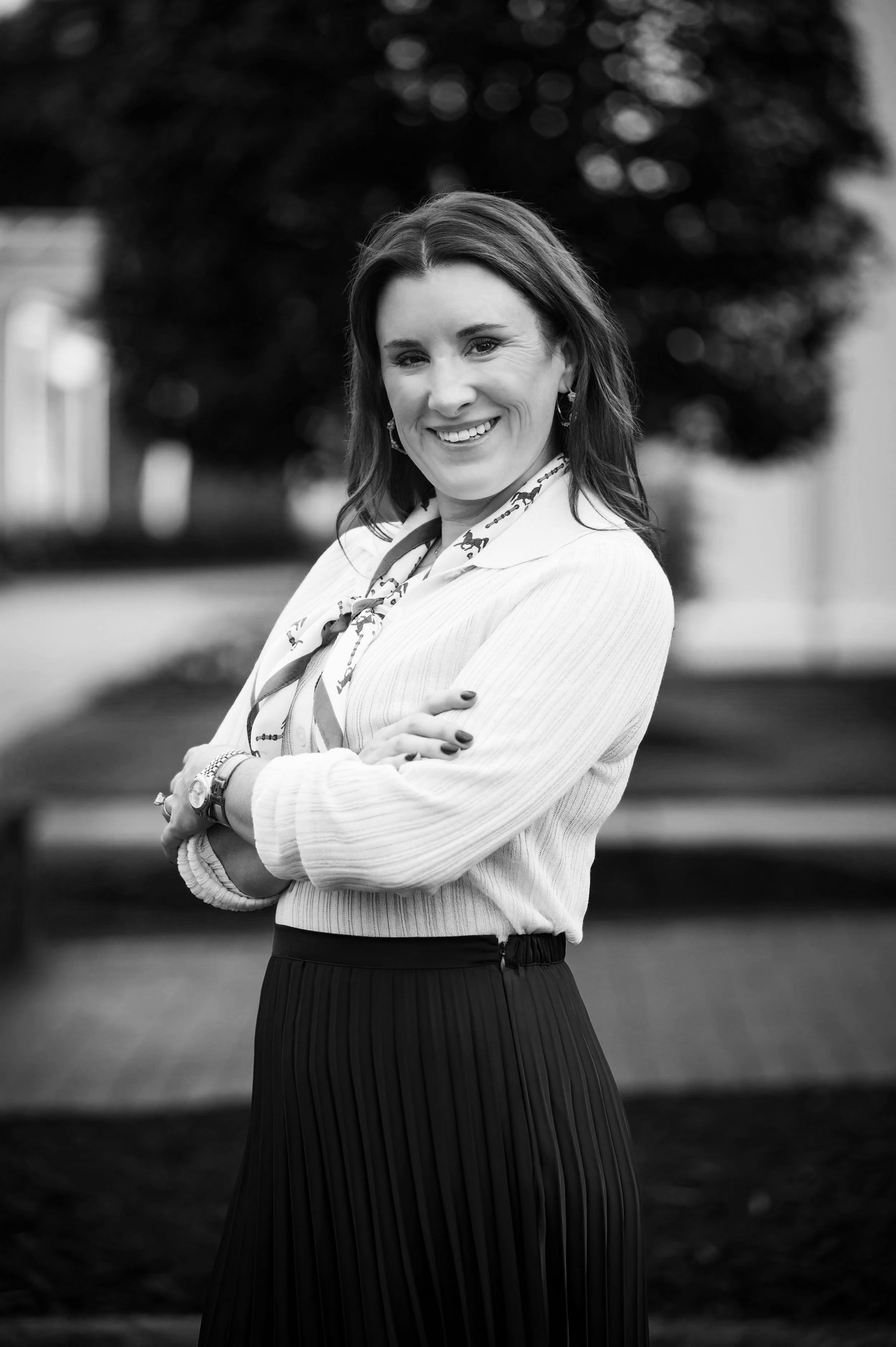 A woman with shoulder-length hair smiling and crossing her arms outdoors, dressed in a light-colored sweater and a dark pleated skirt, with a patterned scarf around her neck.