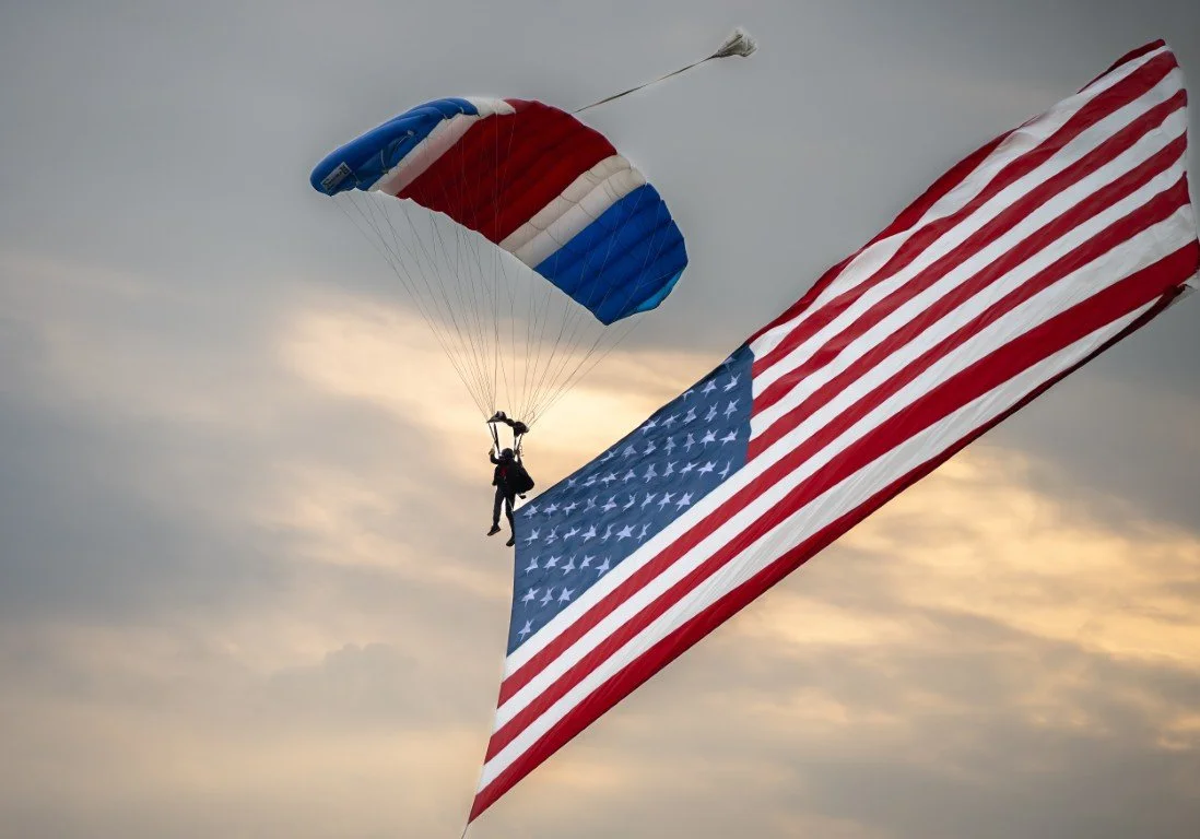 A skydiver holding an American flag while descending with a parachute that has red, white, and blue colors.
