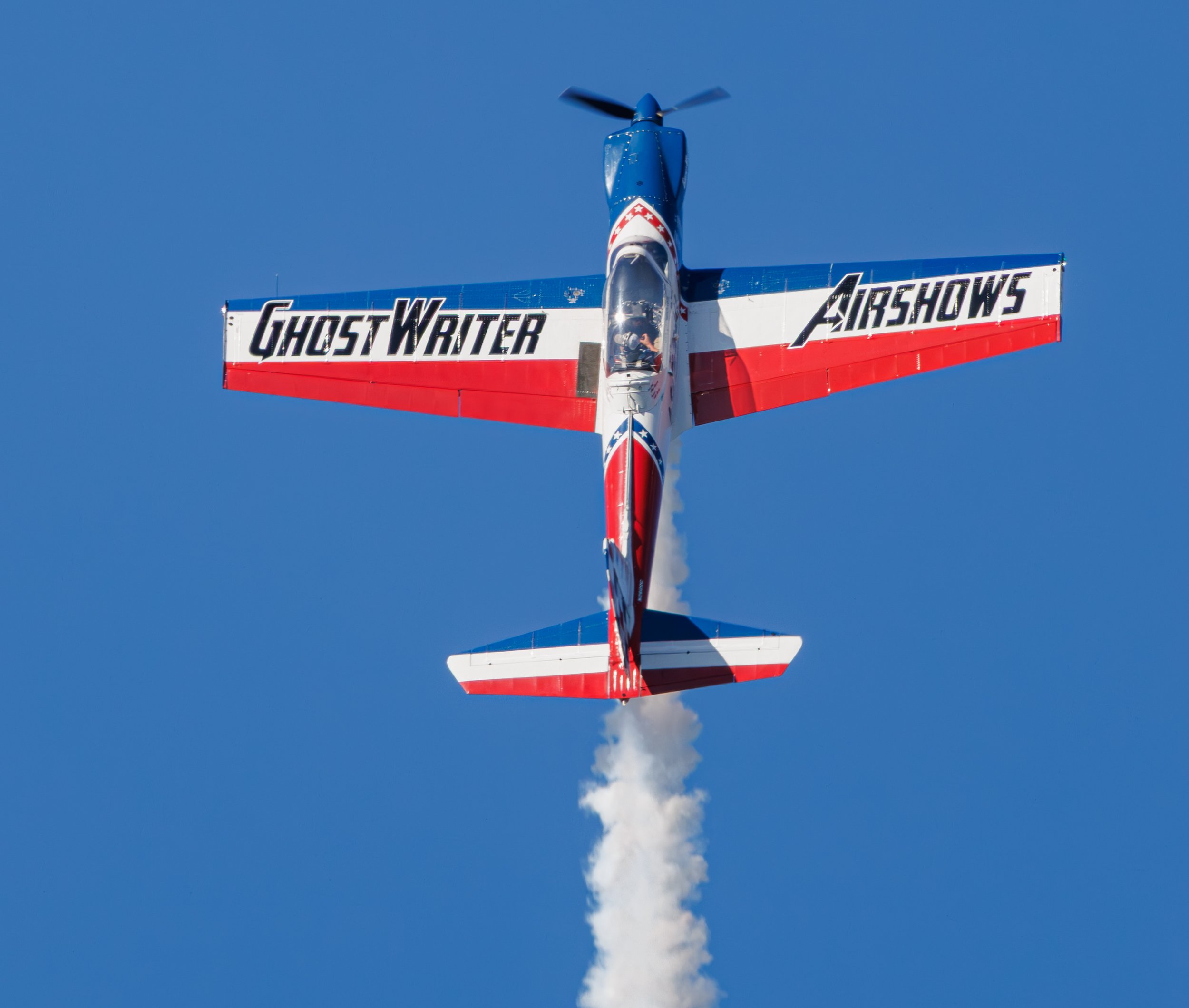 A blue, red, and white stunt plane performing an aerobatic maneuver in the sky, leaving a trail of white smoke. The plane has the words "GhostWriter" on one wing and "AirShows" on the other.