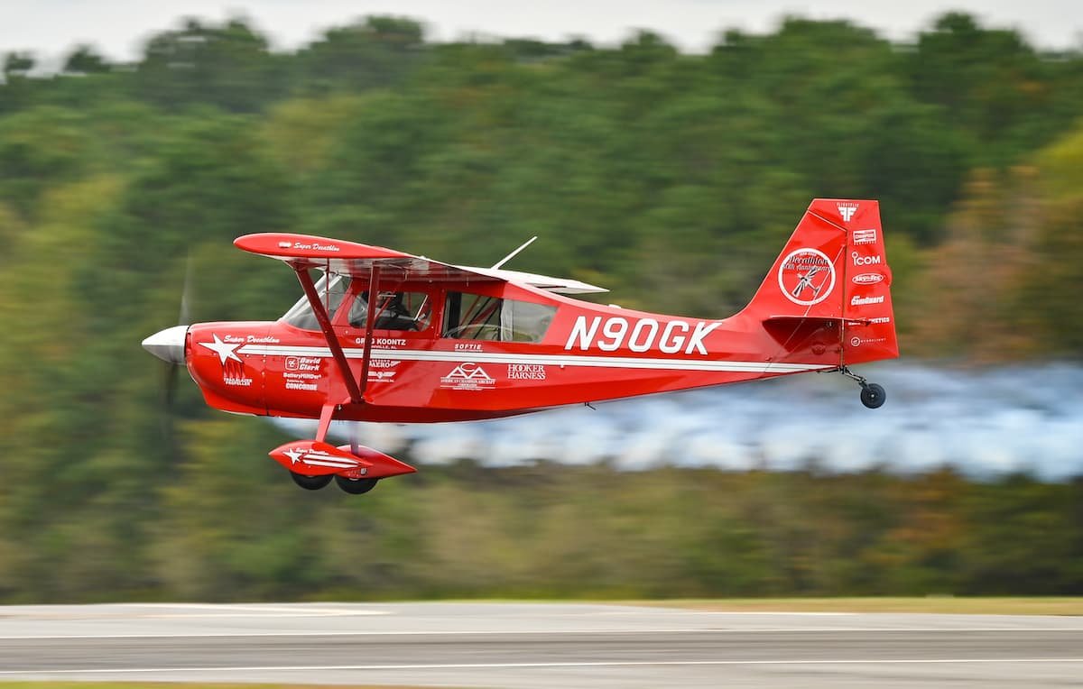 Red aerobatic aircraft flying low over an airstrip with a trail of smoke behind it, surrounded by a wooded landscape.