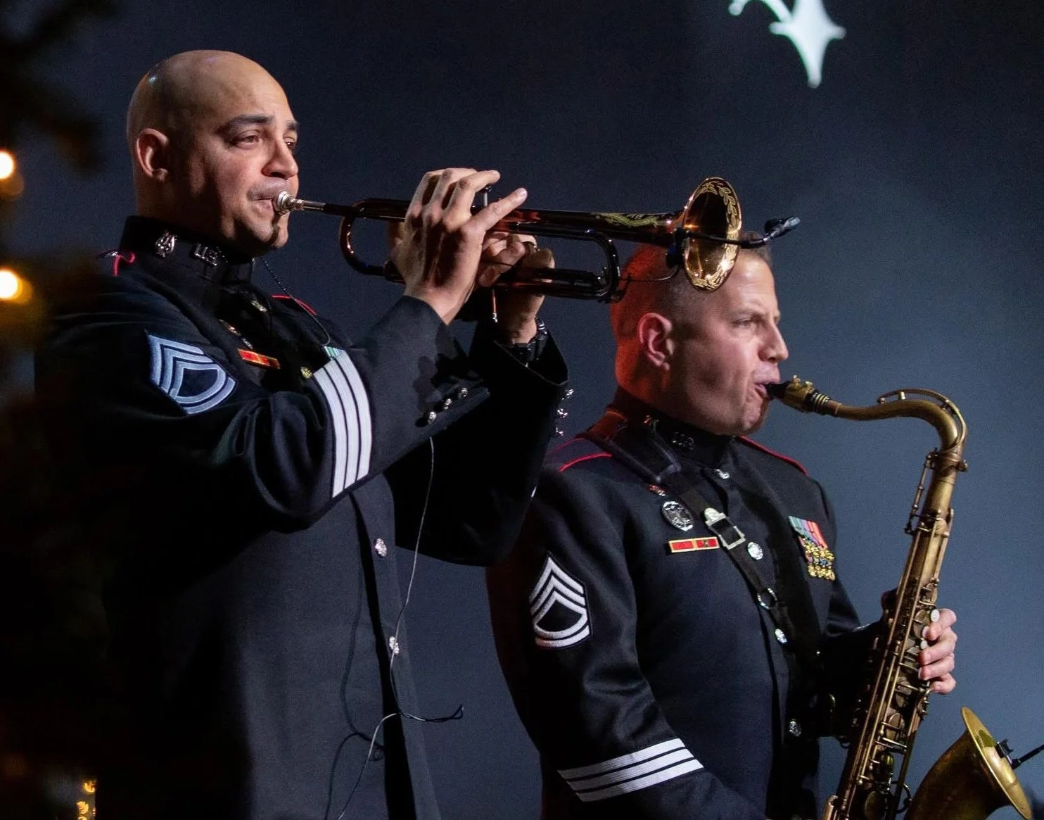 Two military musicians playing brass instruments, with one playing a trumpet and the other playing a saxophone, dressed in formal military uniforms with medals and insignia.