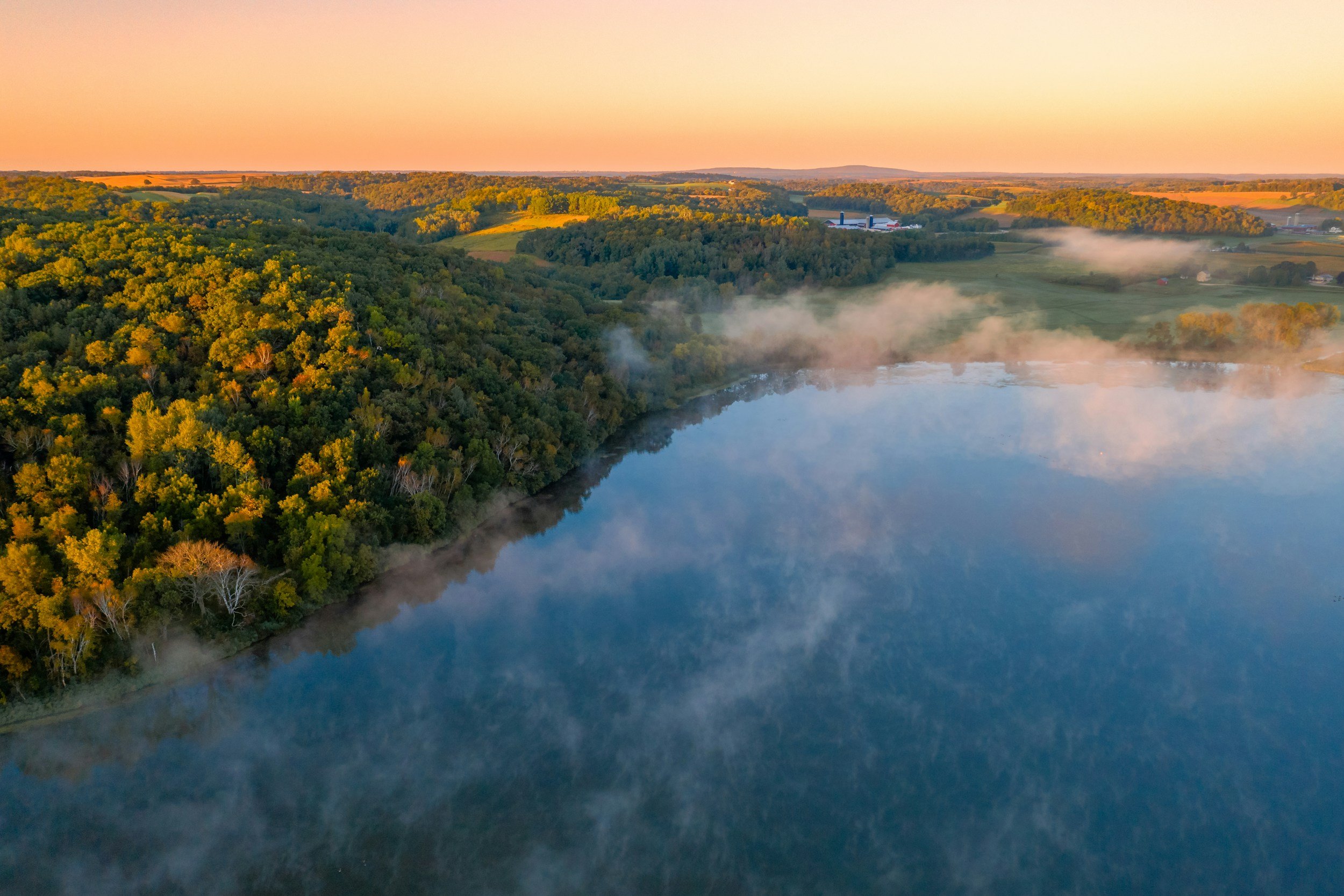 Aerial view of a lake with mist rising from the water, surrounded by green forests and rolling farmland under a pastel sunset sky.