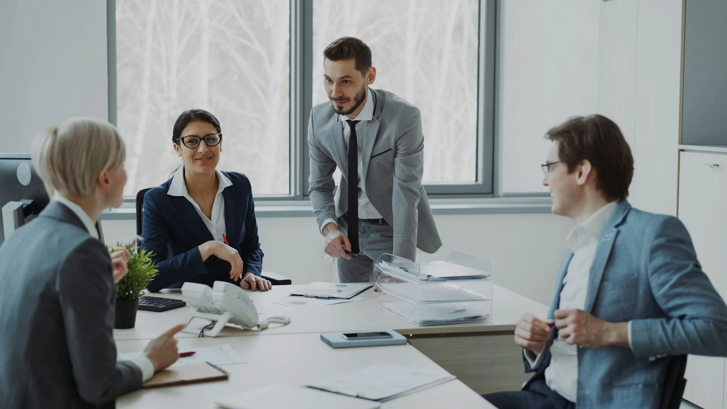 Business meeting with four professionals in a modern office, two women and two men, engaging in discussion around a desk with documents, computers, and office supplies.