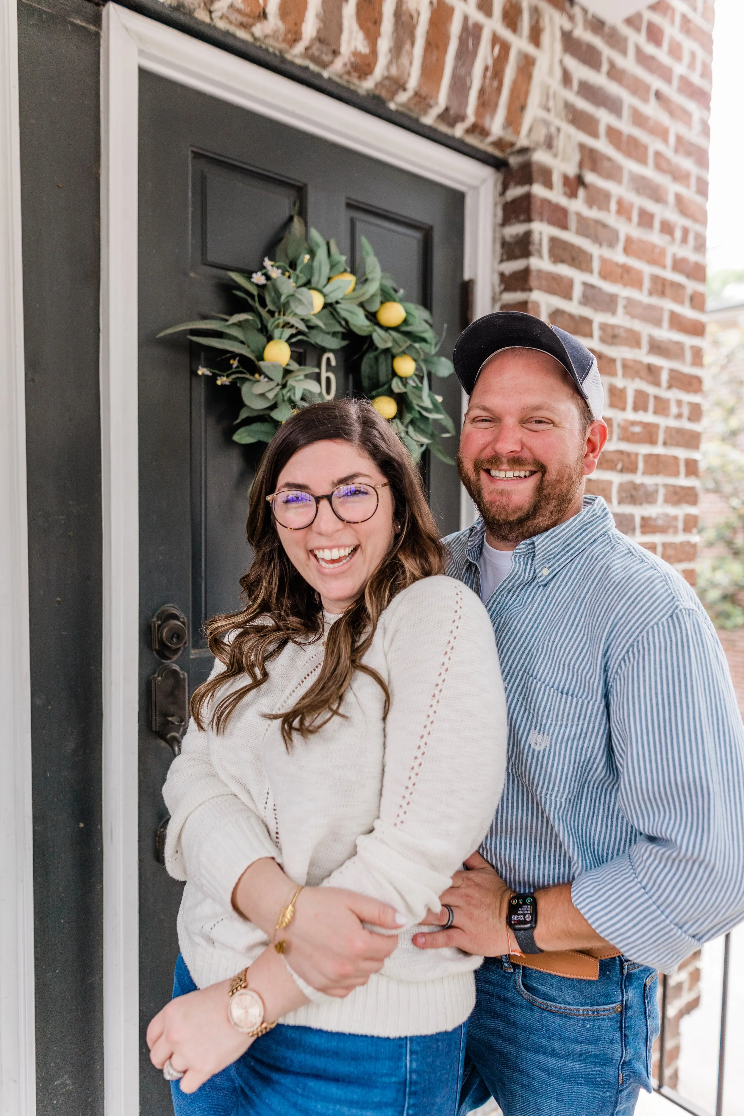 A smiling couple stands in front of a dark gray front door decorated with a lemon and greenery wreath. They are hugging, in savannah georgia historic district lee and alyssa acker of hosting savannah and before and ackers home renovations