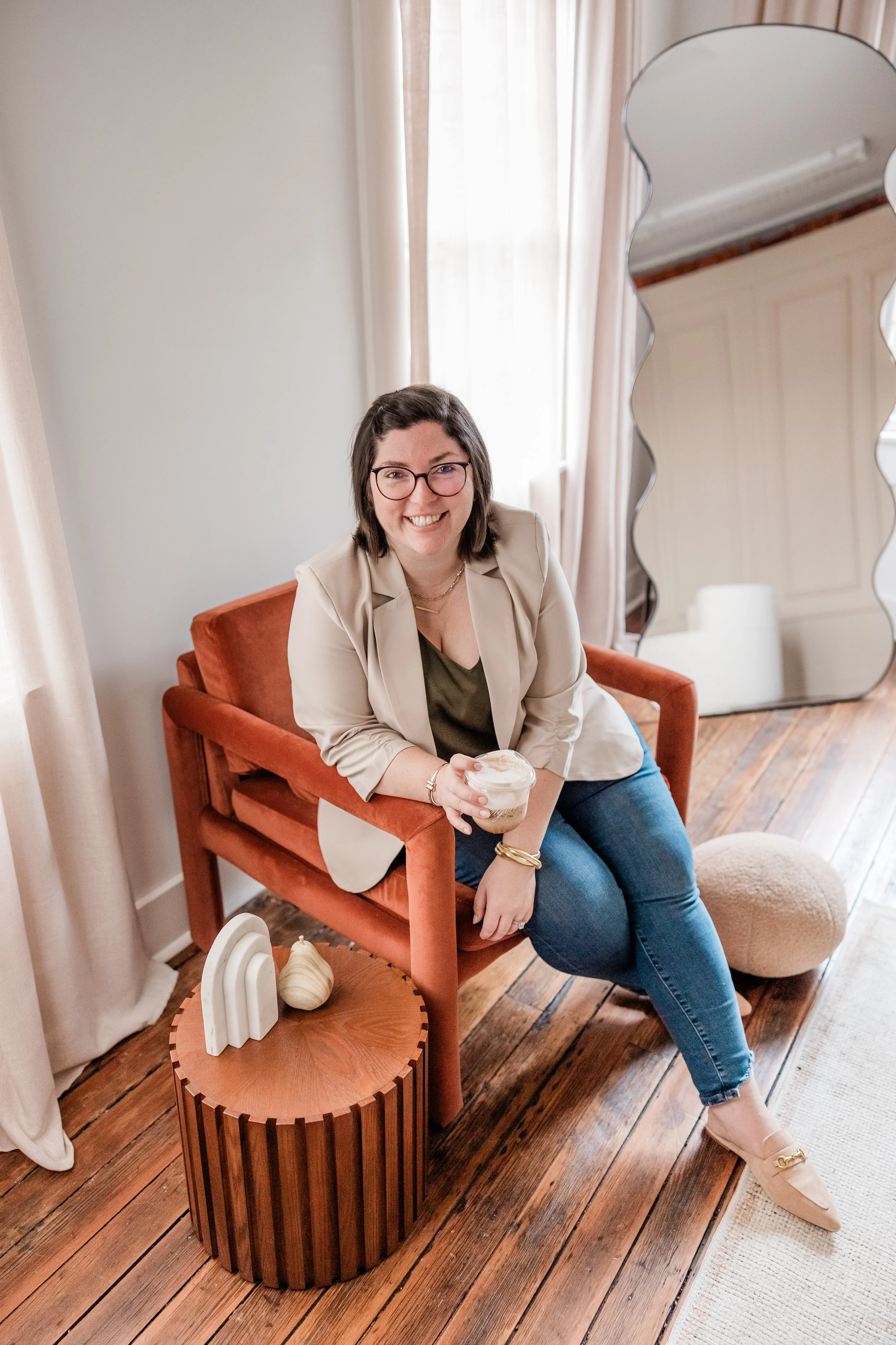 A woman with glasses sitting on an orange chair, holding a beverage, in a room with white curtains, a large mirror, and wooden flooring at wonderland studios in savananh georgia, brandshoot with wendy at Apt B Photo