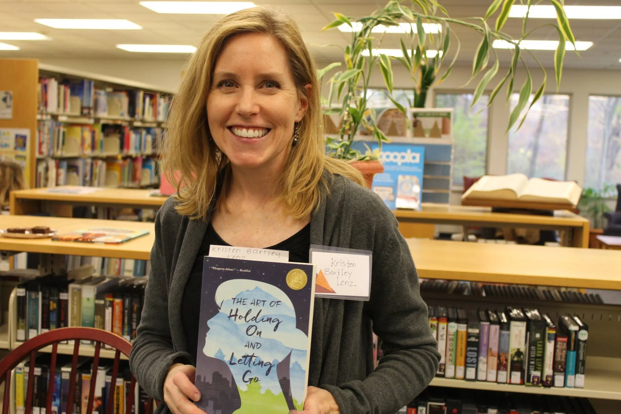 A woman with blond hair smiling while holding a book titled 'The Art of Holding On and Letting Go' in a library.
