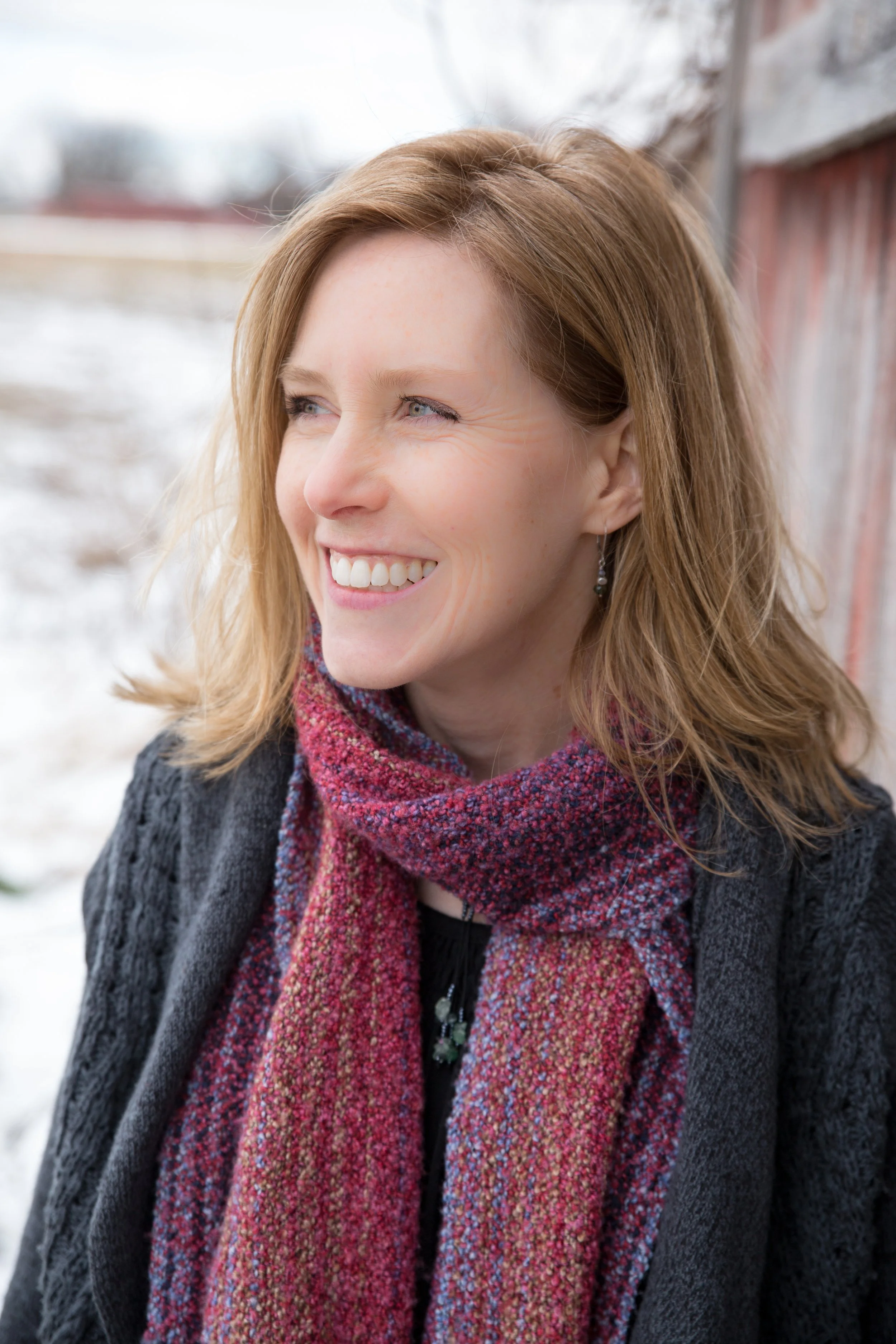 A woman with shoulder-length red hair smiling outdoors near a rustic building, wearing a gray coat and a multicolored scarf.